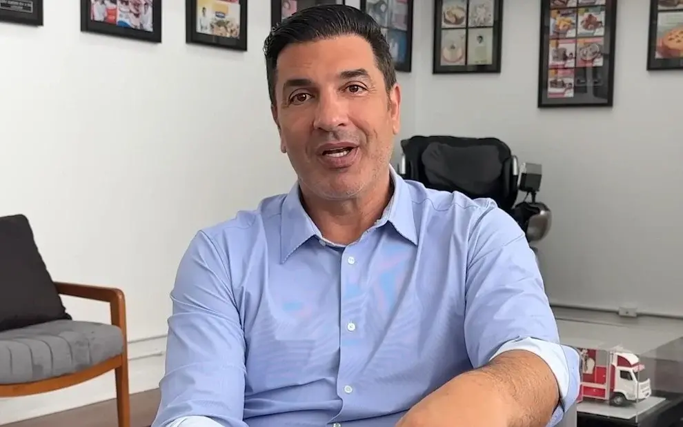 Imagem mostra Edu Guedes falando para a câmera, vestindo uma camisa azul. O fundo da imagem é branco, com alguns quadros na parede.