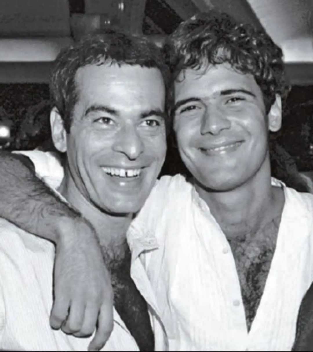 Reprodução de uma foto em preto e branco de dois homens sorrindo e se abraçando, um deles segurando um cigarro, em um ambiente de confraternização.