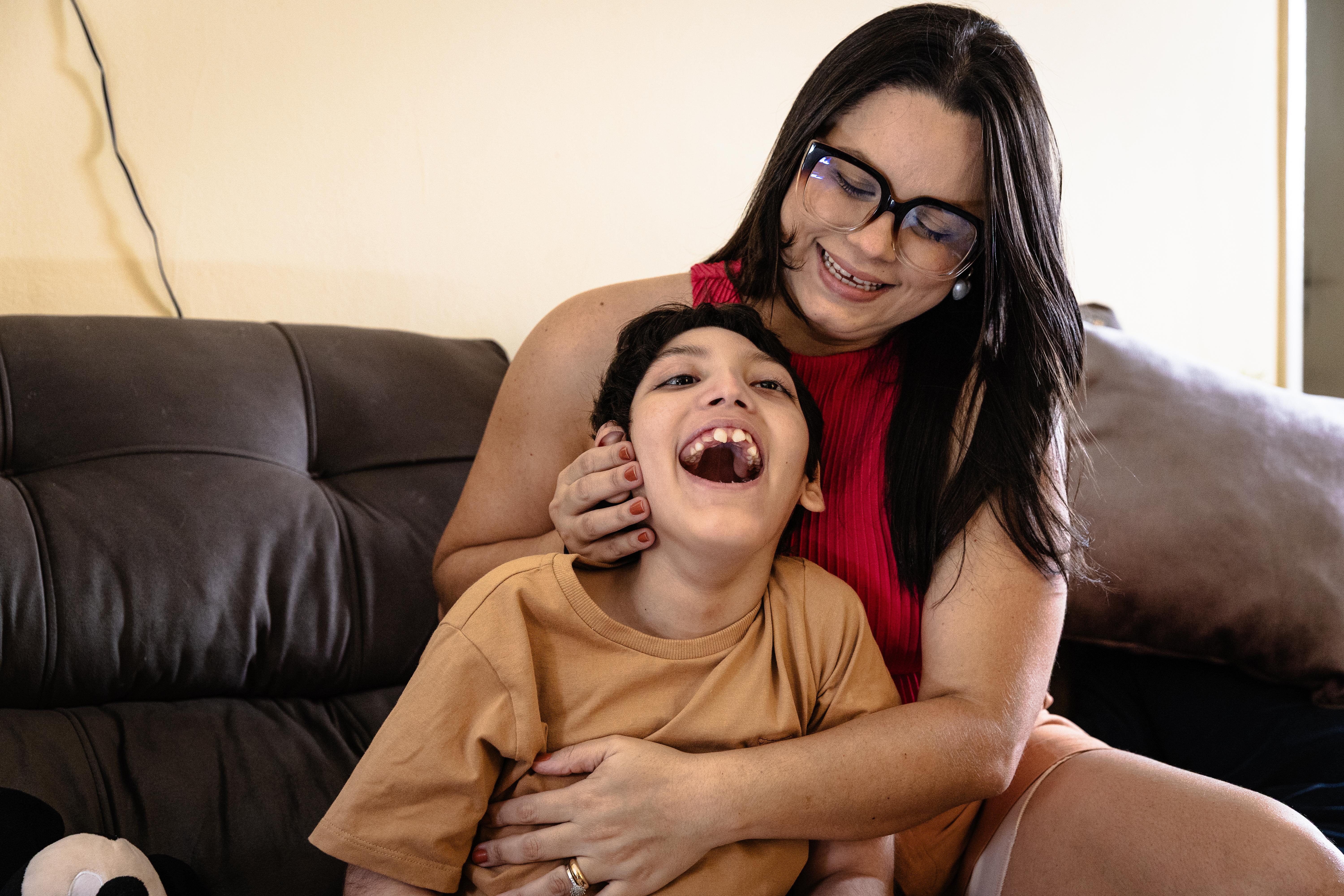 A foto mostra uma jovem com cabelo escuro e óculos, vestindo uma blusa vermelha, está sorrindo e abraçando uma criança com cabelo escuro que veste uma camiseta cor de mostarda. A criança está olhando para cima com a boca aberta, parecendo rir alegremente. Eles estão sentados em um sofá marrom.
