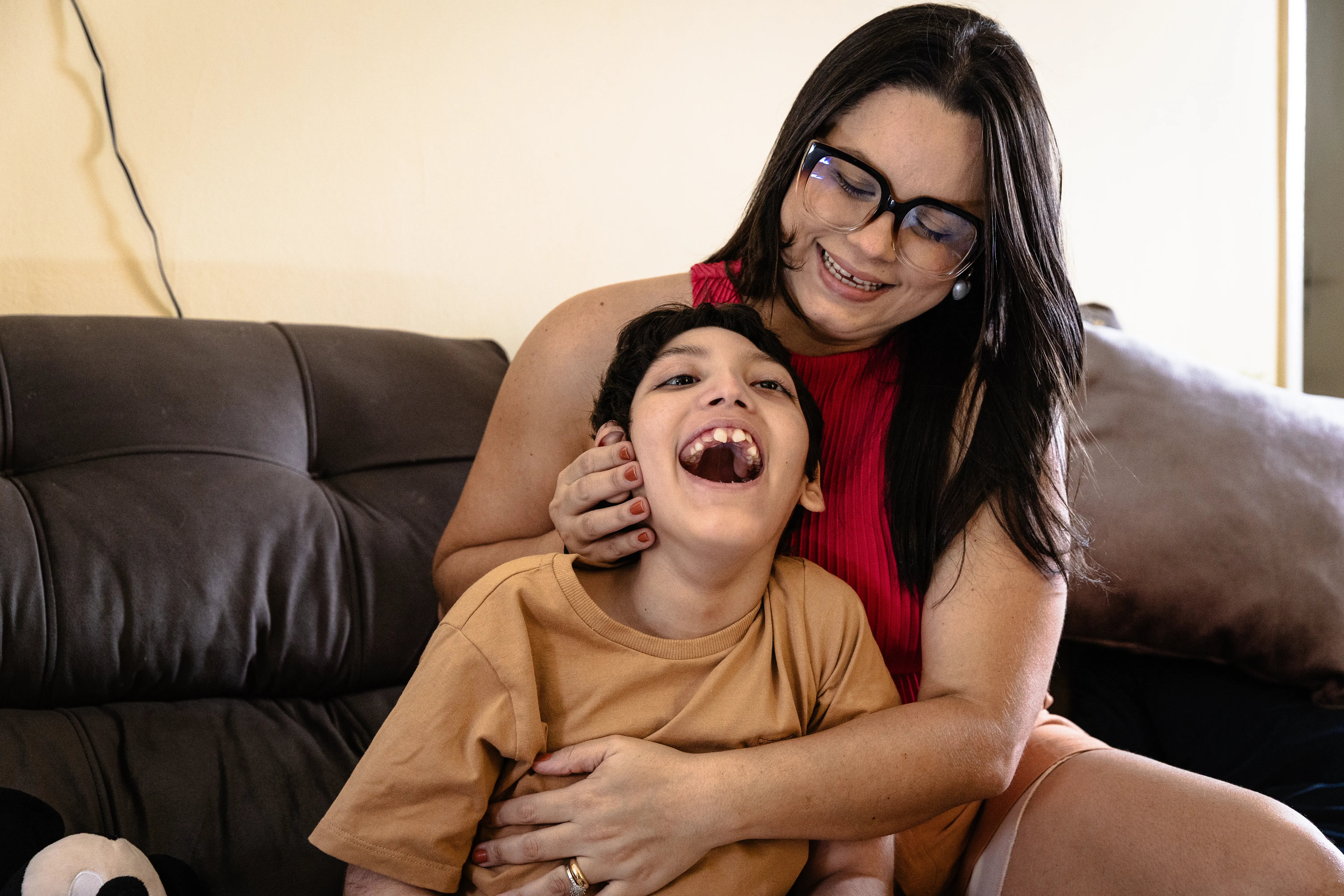 A foto mostra uma jovem com cabelo escuro e óculos, vestindo uma blusa vermelha, está sorrindo e abraçando uma criança com cabelo escuro que veste uma camiseta cor de mostarda. A criança está olhando para cima com a boca aberta, parecendo rir alegremente. Eles estão sentados em um sofá marrom.