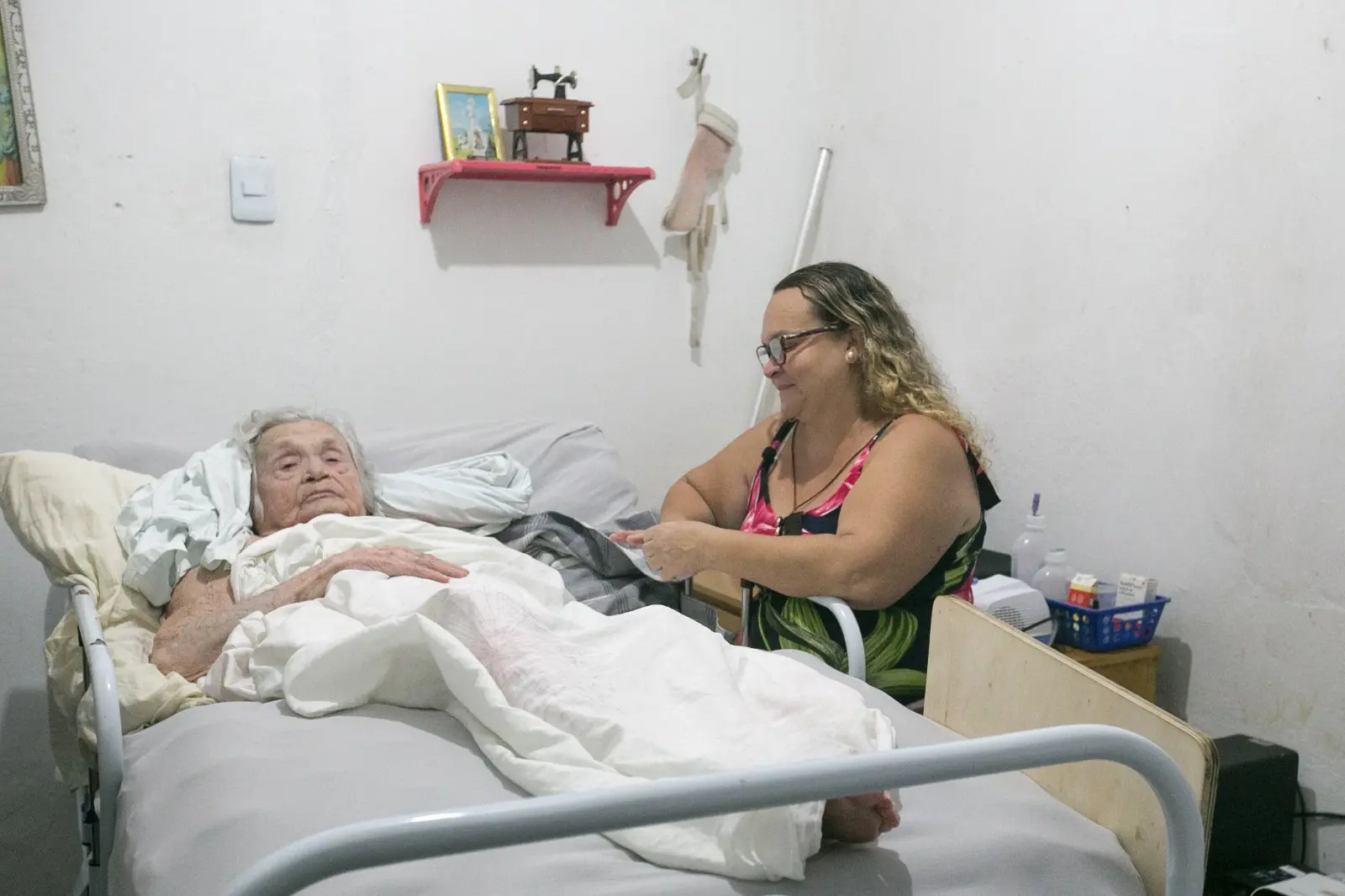 Mulher idosa deitada em uma cama hospitalar, coberta com lençóis brancos, em um quarto simples. Ao lado dela, uma cuidadora sorridente, usando óculos e vestido estampado, segura a mão da idosa com carinho. Ao fundo, há uma prateleira vermelha com objetos decorativos e uma pequena cômoda com remédios e produtos de higiene. O ambiente transmite cuidado e acolhimento.