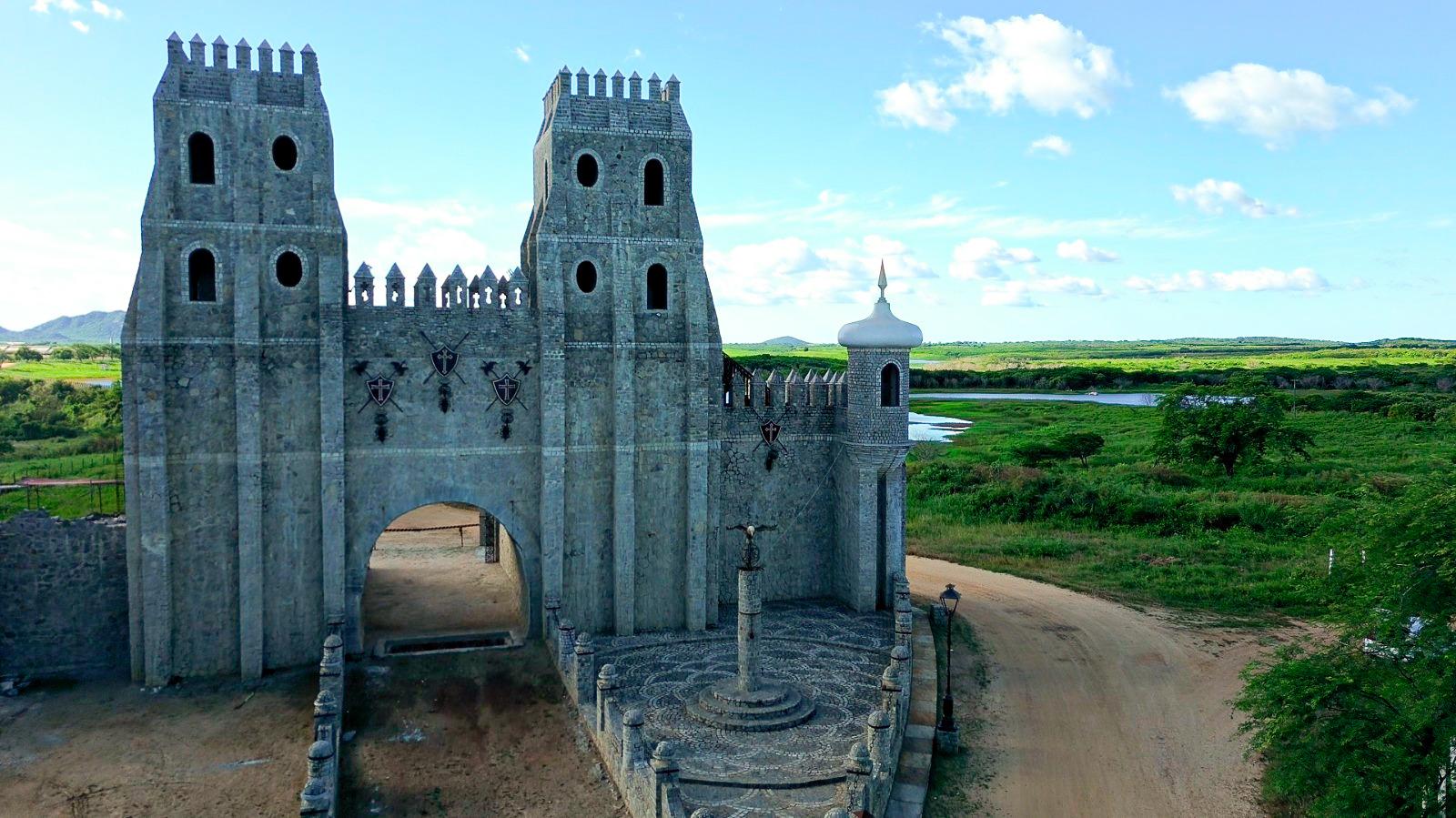 A imagem mostra uma construção de pedra imponente, similar a um castelo medieval, com torres e arco. Ao fundo, natureza e céu.