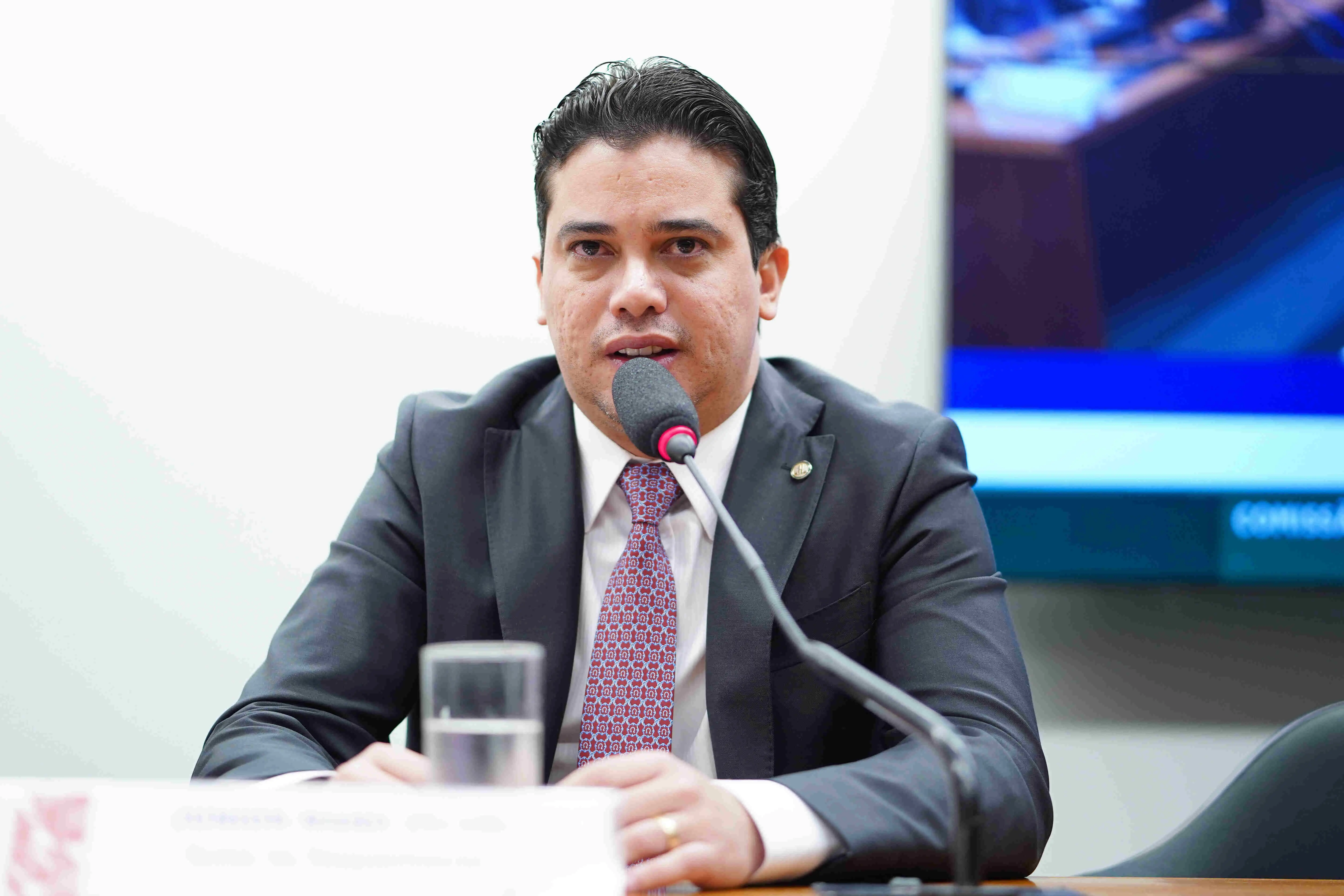 Junior Mano está de terno escuro e gravata vermelha com estampa geométrica e fala ao microfone durante uma audiência ou sessão formal na Câmara dos Deputados. Ele está sentado à mesa, com um copo de água à sua frente e uma televisão ao fundo exibindo parte de um ambiente institucional. A imagem tem aparência oficial, relacionada a um ambiente político.