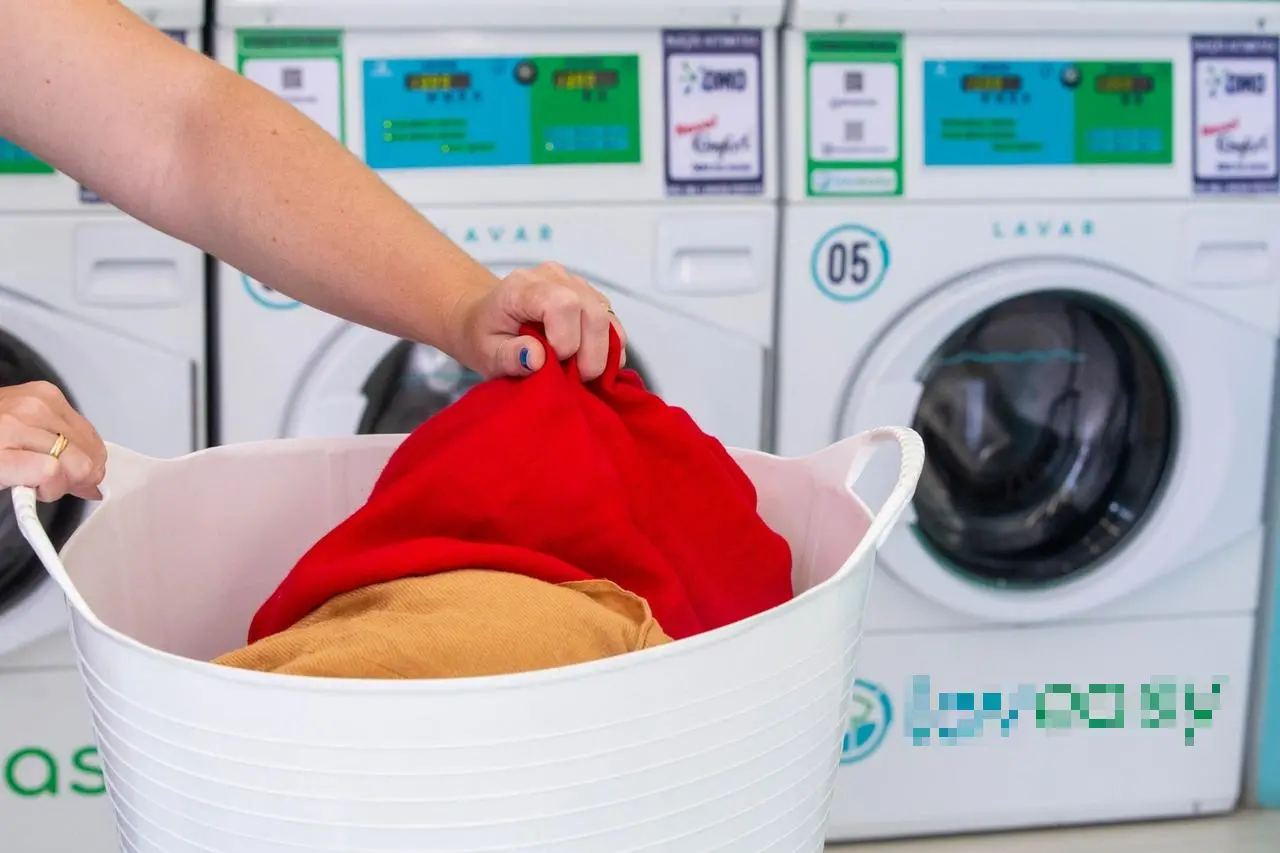 Imagem mostra as mãos de uma mulher branca, que está tirando uma roupa de um cesto em uma lavanderia
