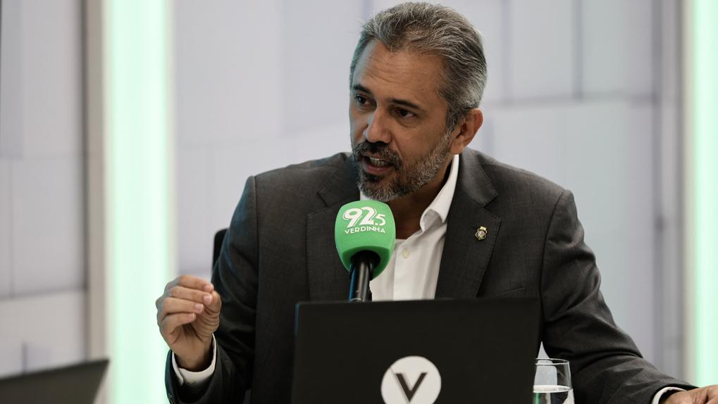 Foto do governador Elmano de Freitas sentado dando entrevista para o programa Bom Dia Nordeste. Ele usa blusa social branca e paletó cinza, com um broche do brasão do Governo do Ceará. À frente dele, há um microfone verde com a logomarca da Verdinha FM e um notebook.