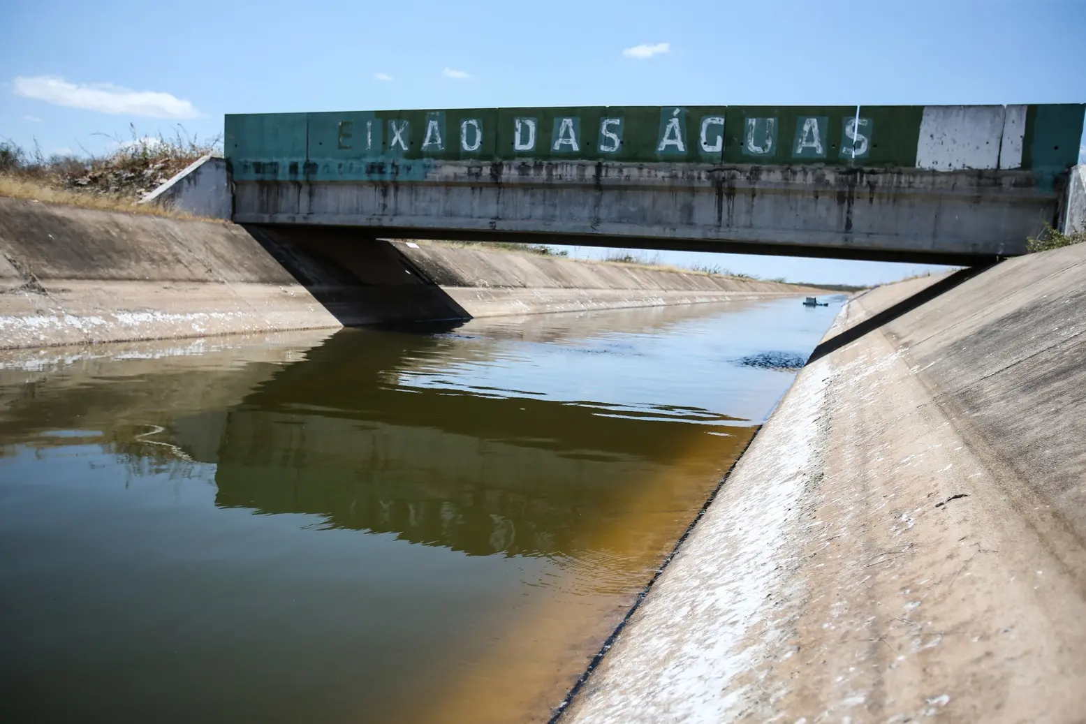 Foto de canal de concreto com água esverdeada, próximo a uma ponte com o letreiro Eixão das Águas, em dia de céu claro com poucas nuvens