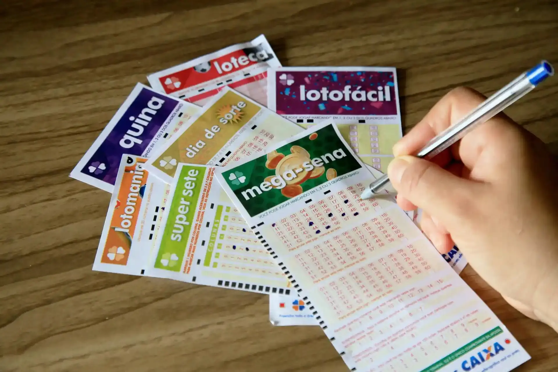 Pessoa marcando números em várias cartelas de jogos de azar das Loterias da Caixa Econômica Federal, como Lotomania, Mega-Sena, Quina, Lotofácil, entre outros, sobre uma mesa de madeira. Novos valores de apostas das Loterias Caixa passam a valer a partir desta quarta (9).