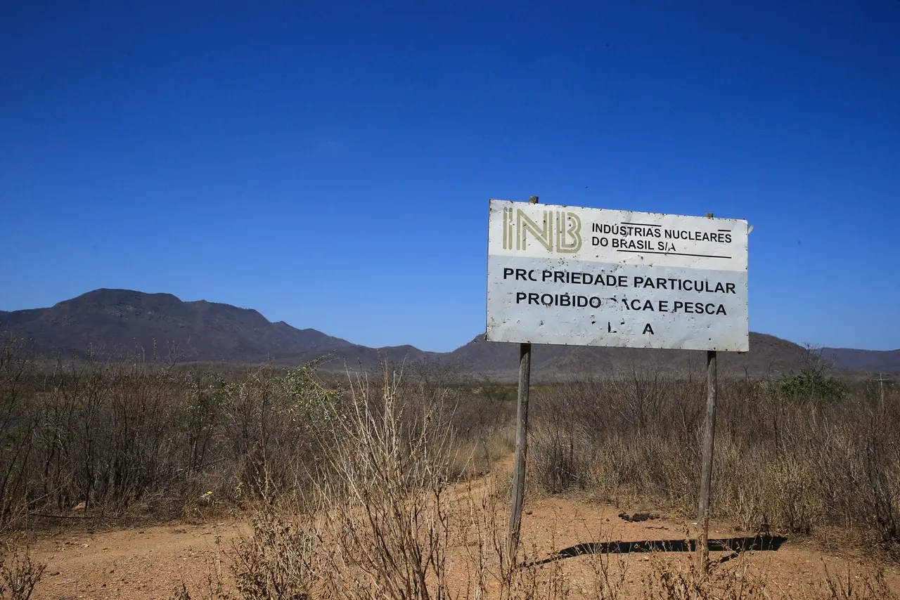 Foto de uma placa com o nome Indústrias Nucleares do Brasil, sendo propriedade particular com proibição de ultrapassar. Paisagem mostra plantas de galhos secos e solo arenoso na cidade de Santa Quitéria