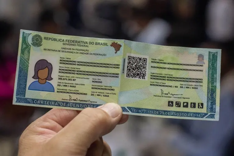A foto mostra uma mão segura um novo modelo de carteira de identidade brasileira, que é verde e amarela e tem um QR code no lado direito.