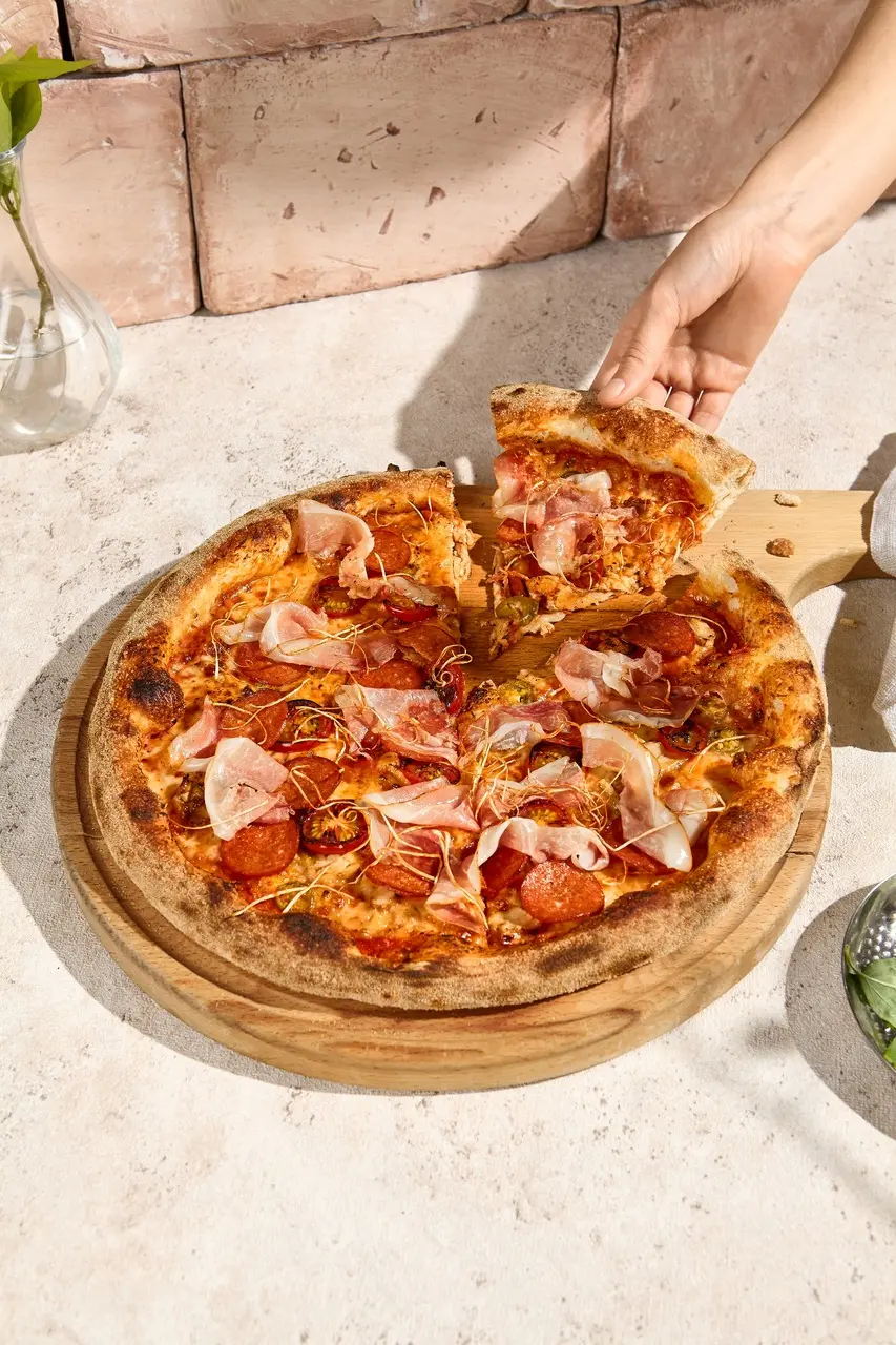 Pizza de borda grossa coberta com pepperoni, presunto, queijo mussarela e almôndegas, servida em uma tábua de madeira, pronta para ser apreciada, ilustrando receita de massa de pizza no liquidificador no dia nacional da pizza.