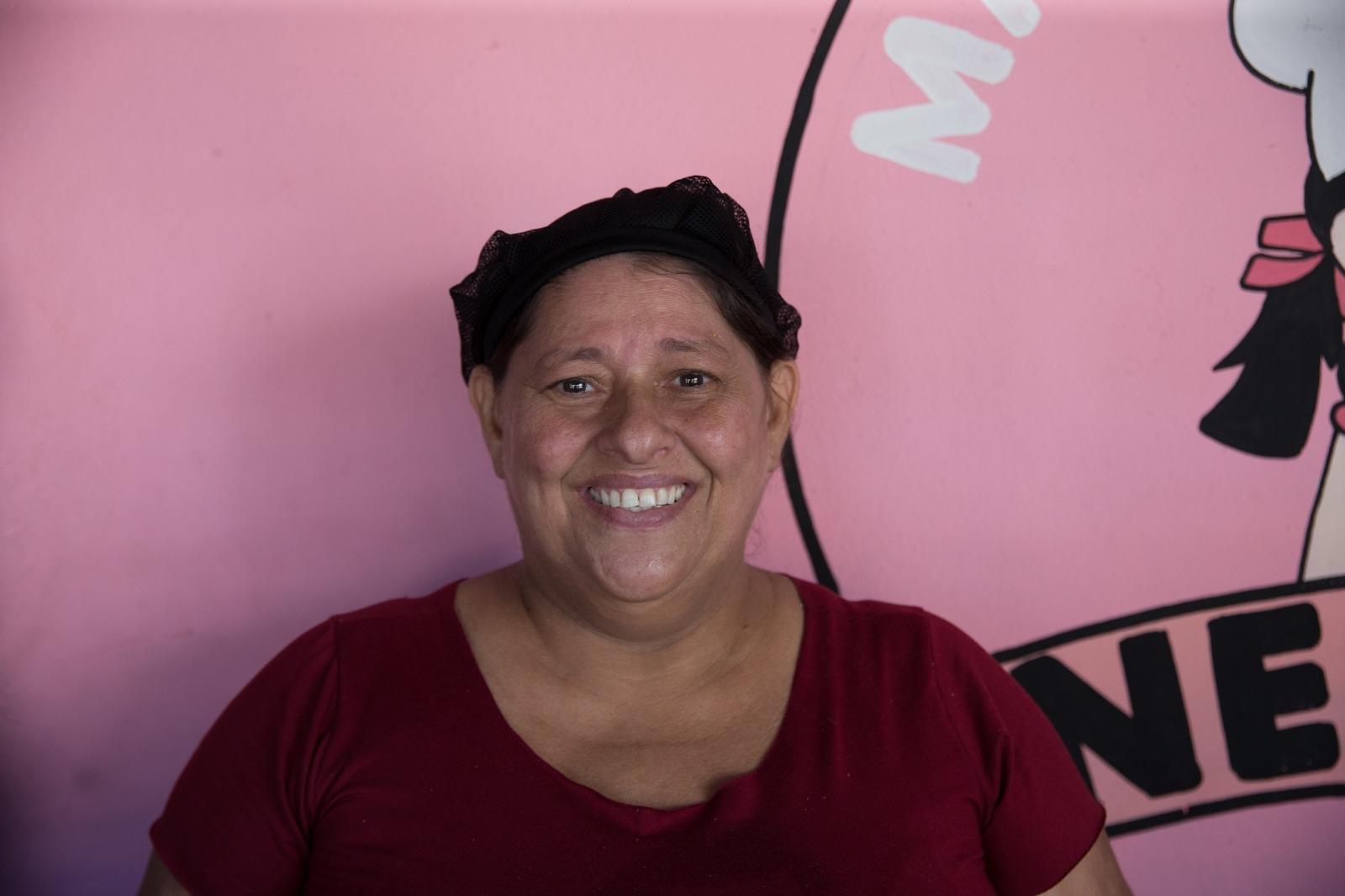 Imagem mostra mulher de touca preta e camisa cor de vinho sorrindo. Ao fundo, uma parede rosa.