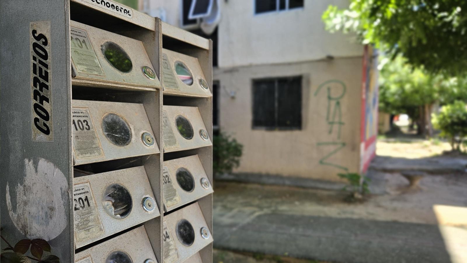 Imagem mostra, em primeiro plano, caixas de correio de cor cinza, com números de apartamentos. Ao fundo, uma parede está pichada com a palavra Paz.
