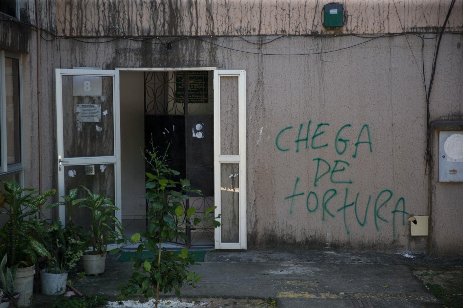 Imagem mostra a entrada de um dos blocos do residencial José Euclides, em Fortaleza. À esquerda da foto, uma porta de vidro com moldura de metal está aberta. À direita, no muro, há uma pichação com a frase chega de tortura.
