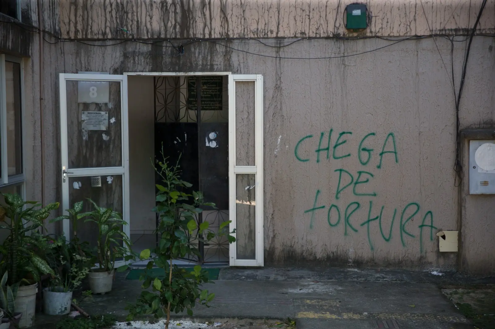 Imagem mostra a entrada de um dos blocos do residencial José Euclides, em Fortaleza. À esquerda da foto, uma porta de vidro com moldura de metal está aberta. À direita, no muro, há uma pichação com a frase chega de tortura.