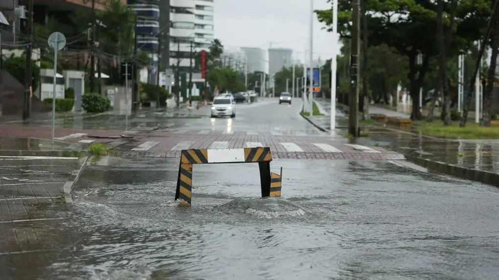 A foto mostra uma rua urbana inundada pela chuva, com um bueiro transbordando em primeiro plano, sinalizado por uma barreira preta e amarela. Ao fundo, carros trafegam pela via alagada, ladeada por prédios altos e árvores. O céu está nublado, indicando um dia chuvoso.