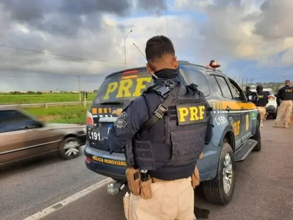Policial da PRF ao lado de um carro da polícia rodoviária federal durante operação na estrada, com outros policiais ao fundo e céu com nuvens, em cenário de fiscalização de trânsito.