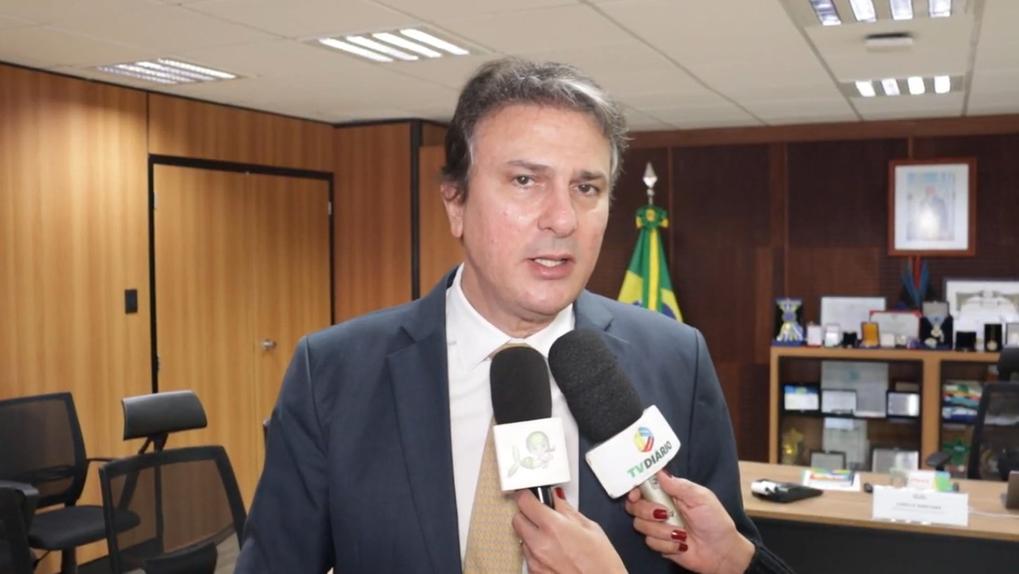 Camilo usa terno azul-marinho, camisa branca e gravata bege, e aparece em uma sala de escritório, falando à imprensa. Ele está sendo entrevistado por dois microfones, um com o logotipo da TV Diário e outro com símbolo da TV Verdes Mares. Ao fundo, há uma estante com objetos de decoração, troféus e documentos, além de uma bandeira do Brasil. As paredes são revestidas de madeira clara e o teto possui iluminação fluorescente.