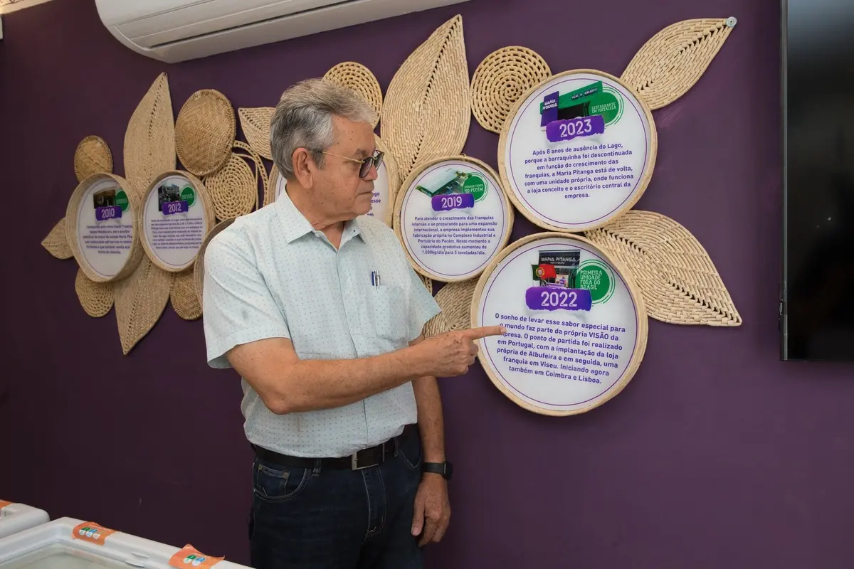 Personagem mostra um mural com a trajetória dele. Homem branco e grisalho