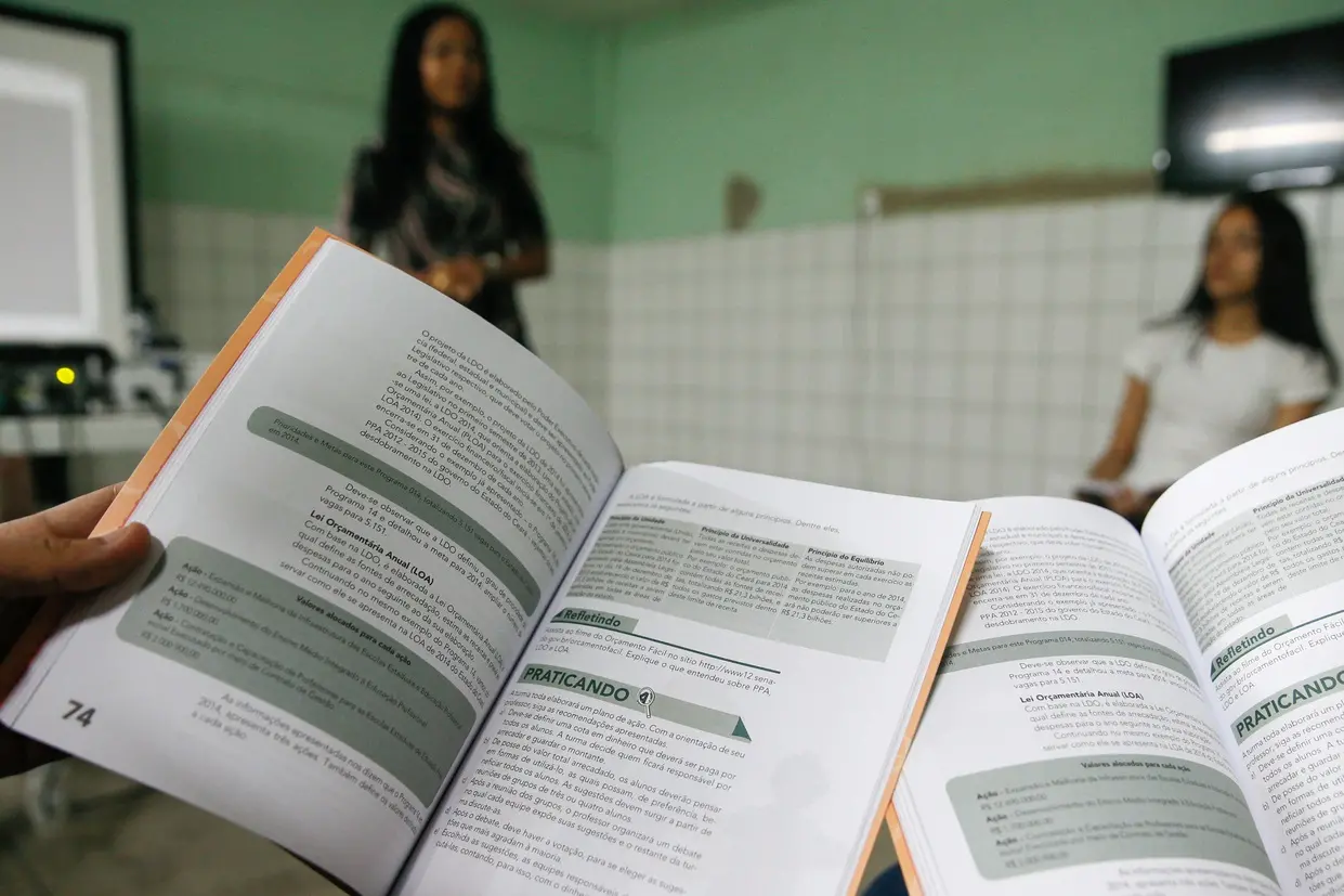 Foto mostra um livro aberto em primeiro plano com conteúdo didático, enquanto ao fundo duas alunas participam de uma atividade em sala de aula. O ambiente é escolar com paredes azulejadas brancas.