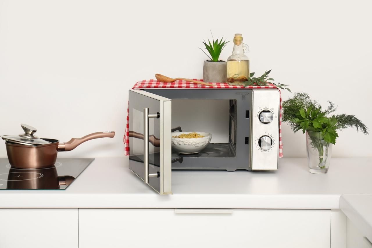 Imagem de micro-ondas na cozinha com alimentos, plantas e utensílios domésticos ao redor, ilustrando se aquecer comida no eletrodoméstico é seguro ou não