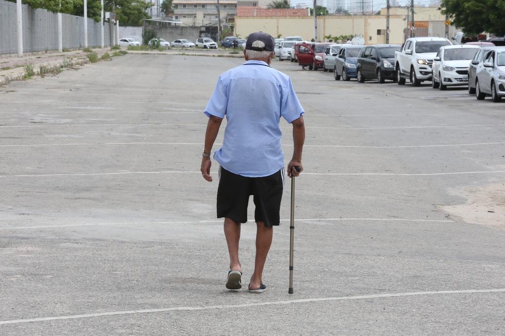 A imagem mostra um idoso de costas, usando chapéu, camisa azul e bermuda preta, caminhando com uma bengala em um estacionamento vazio com carros estacionados ao longe.