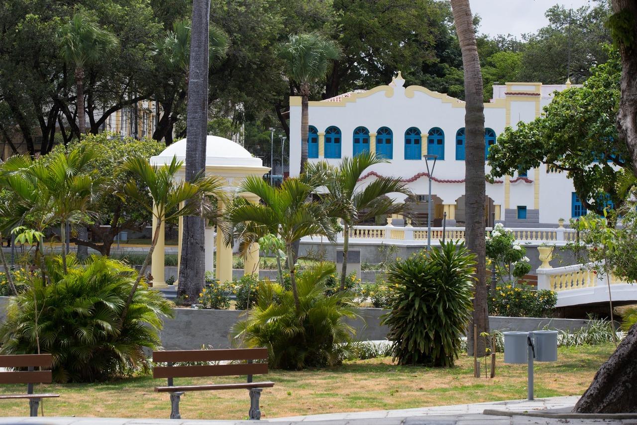 Foto de parque em Fortaleza, a cidade da Criança, com prédio antigo pintado em branco e amarelo, árvores e arbustos verdes e banquinhos