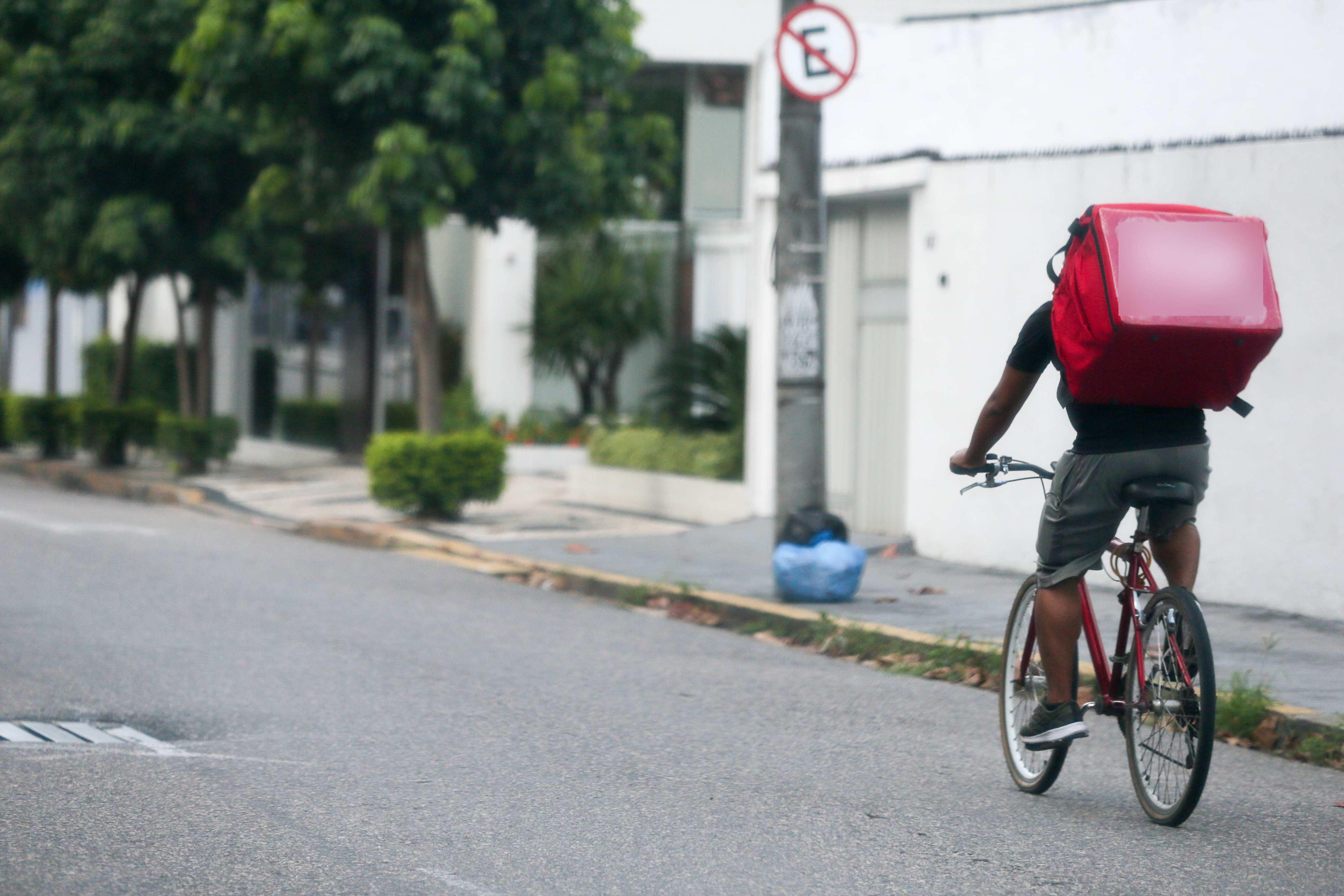 A foto mostra entregador de delivery pedala bicicleta com mochila vermelha em rua arborizada com placa de tr&acirc;nsito.