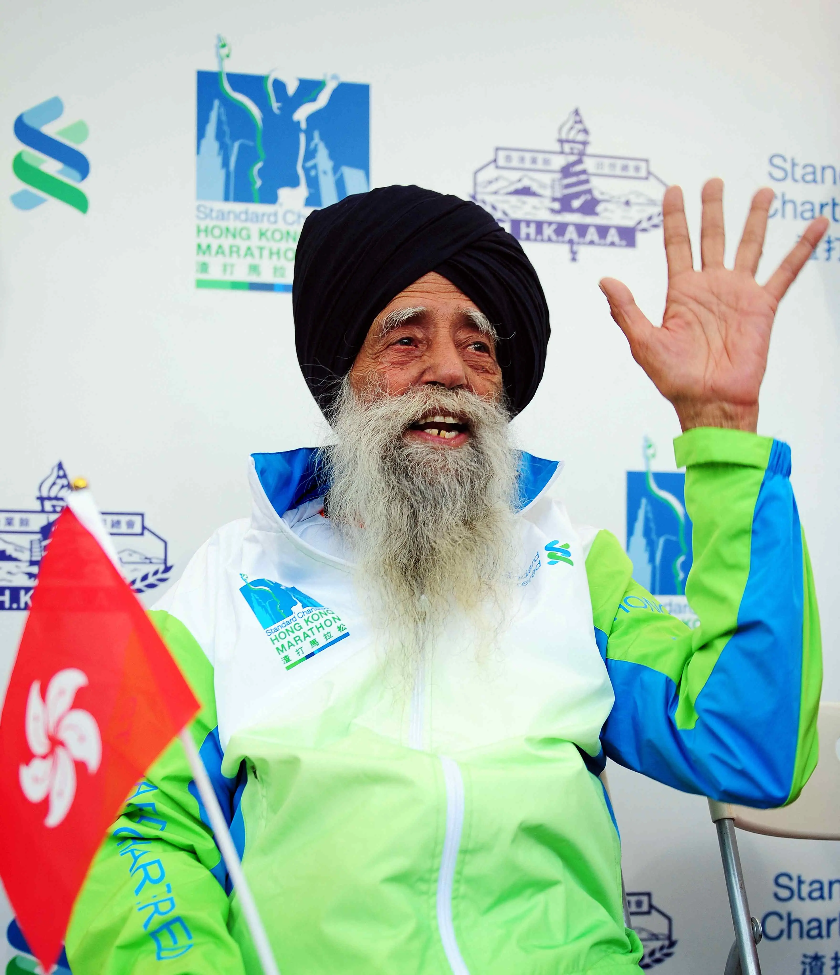 Idoso sorridente com barba branca, usando turbante preto e jaqueta de corrida nas cores verde, azul e branca, acenando na linha de chegada de uma maratona em Hong Kong, com logos de maratona ao fundo.