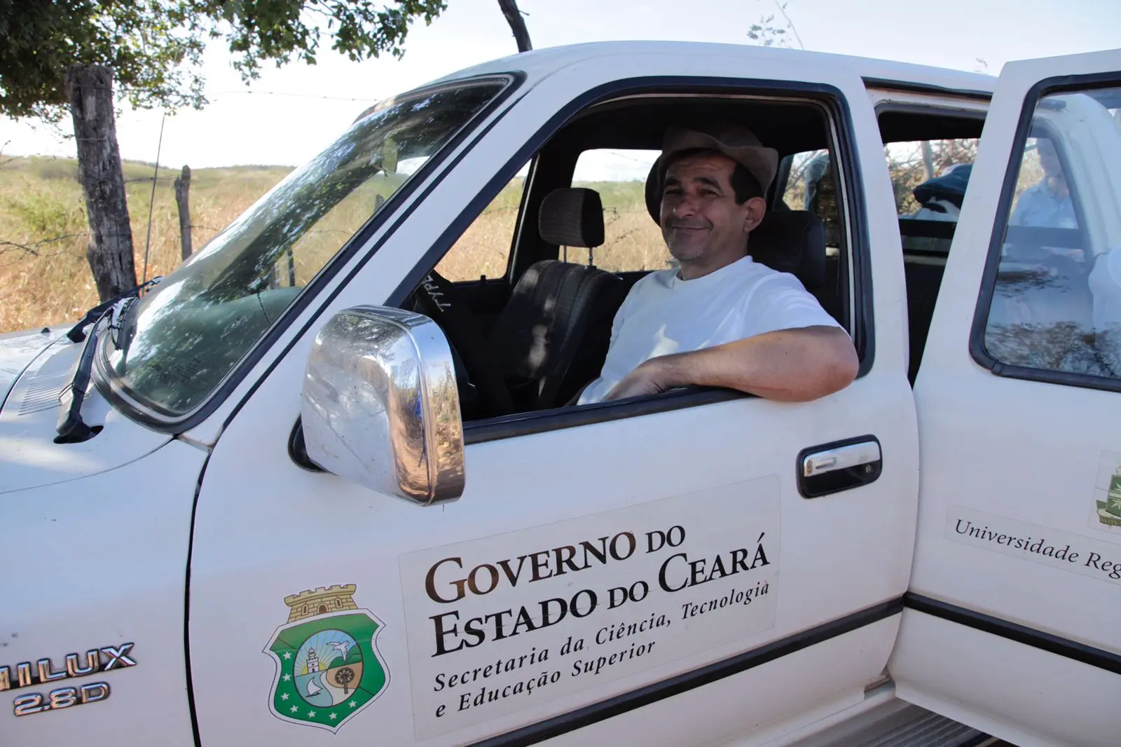 Foto do professor Álamo Saraiva sorridente, usando um chapéu, sentado ao volante de uma caminhonete Hilux do Governo do Estado do Ceará, usada em atividades da Secretaria da Ciência, Tecnologia e Educação Superior. Ao fundo, vê-se uma paisagem rural com vegetação seca.