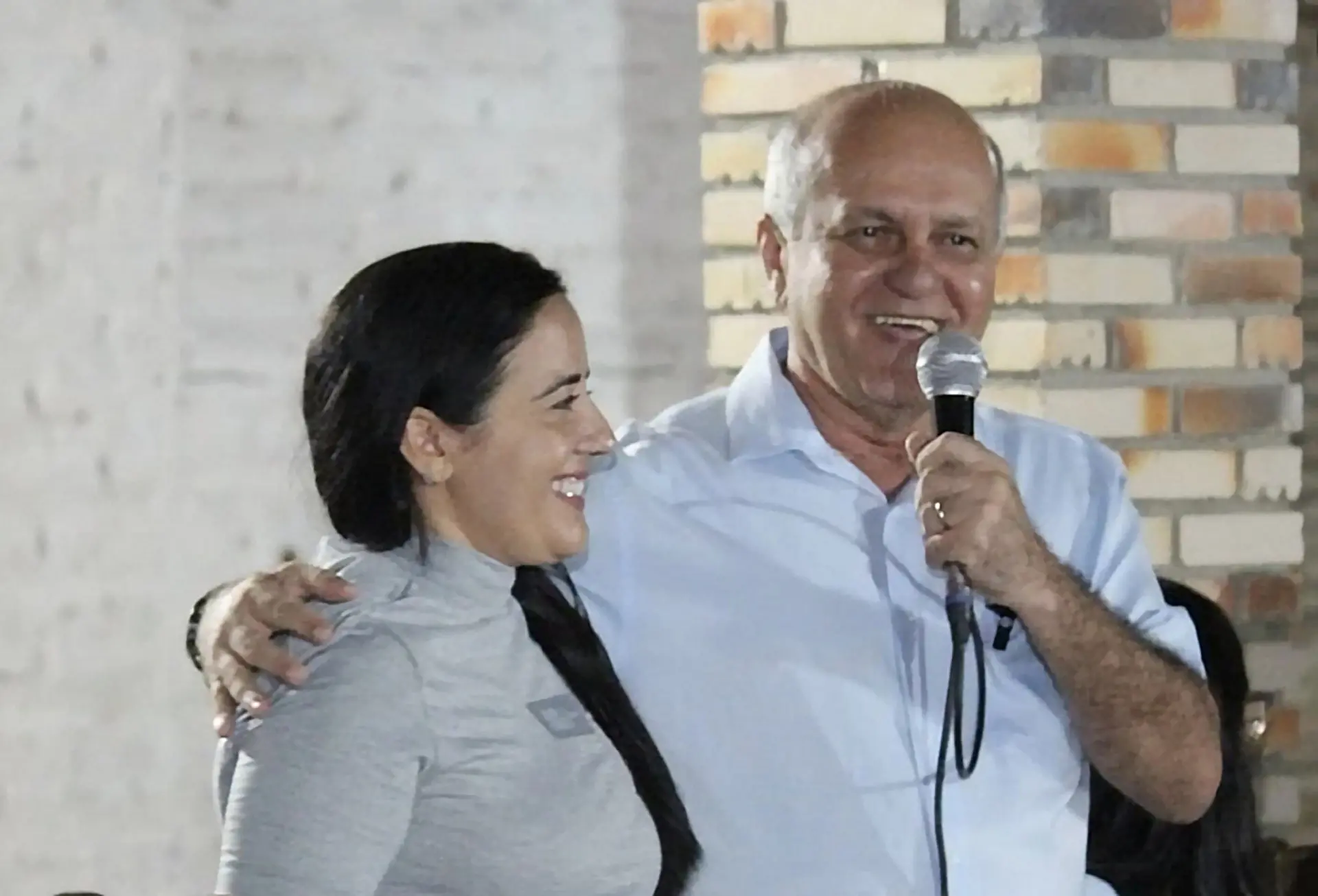 Jaime Veras e Carmen Lúcia sorrindo durante evento em ambiente interno. O homem, de cabelos grisalhos e camisa social azul-clara, segura um microfone e fala enquanto abraça a mulher ao seu lado, que veste uma blusa cinza de manga longa e tem cabelo escuro preso em uma trança. Ambos parecem descontraídos e felizes. Ao fundo, há uma parede de tijolos aparentes. A cena transmite um momento de confraternização ou fala pública em clima amistoso.