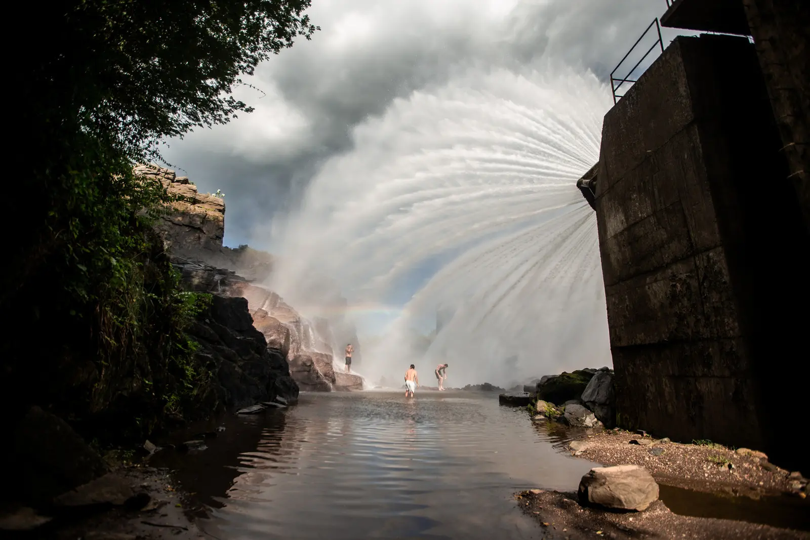 Imagem mostra válvula do açude Orós jorrando água e formando um arco-íris. Ao longe, três homens tomam banho no açude vestindo shorts, sem camisa.