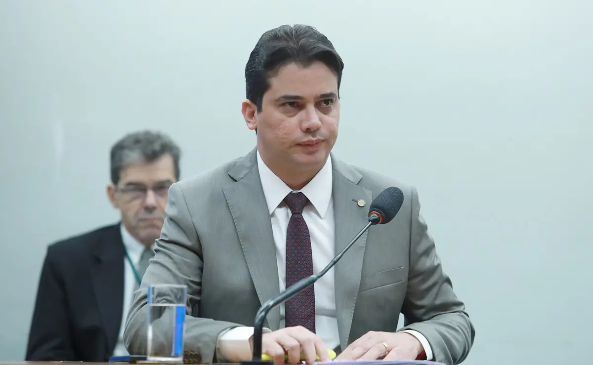 Júnior Mano, homem jovem de terno cinza e gravata marrom, está sentado em uma bancada parlamentar, com expressão séria e olhar voltado para o lado. Ele está em um ambiente institucional, na Câmara dos Deputados, com microfones e outros parlamentares ao fundo. A cena transmite um momento de sessão ou reunião formal.