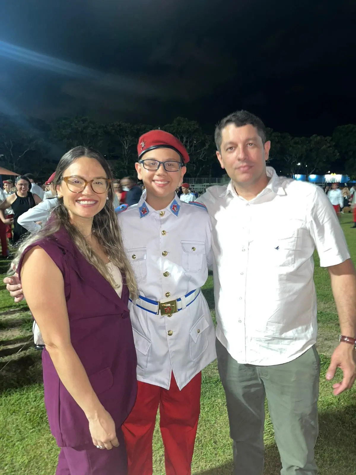 Uma fotografia de uma família de três pessoas, tirada à noite em um gramado. No centro, um jovem sorridente, com óculos, veste um uniforme militar branco com detalhes azuis e dourados, calças vermelhas e um gorro vermelho. Ele está ladeado por uma mulher à sua esquerda e um homem à sua direita. A mulher, com cabelo comprido e óculos, usa um colete cor de vinho sobre uma blusa clara. O homem tem cabelo escuro e veste uma camisa social branca de mangas curtas e calças cinzas. Todos sorriem para a câmera.