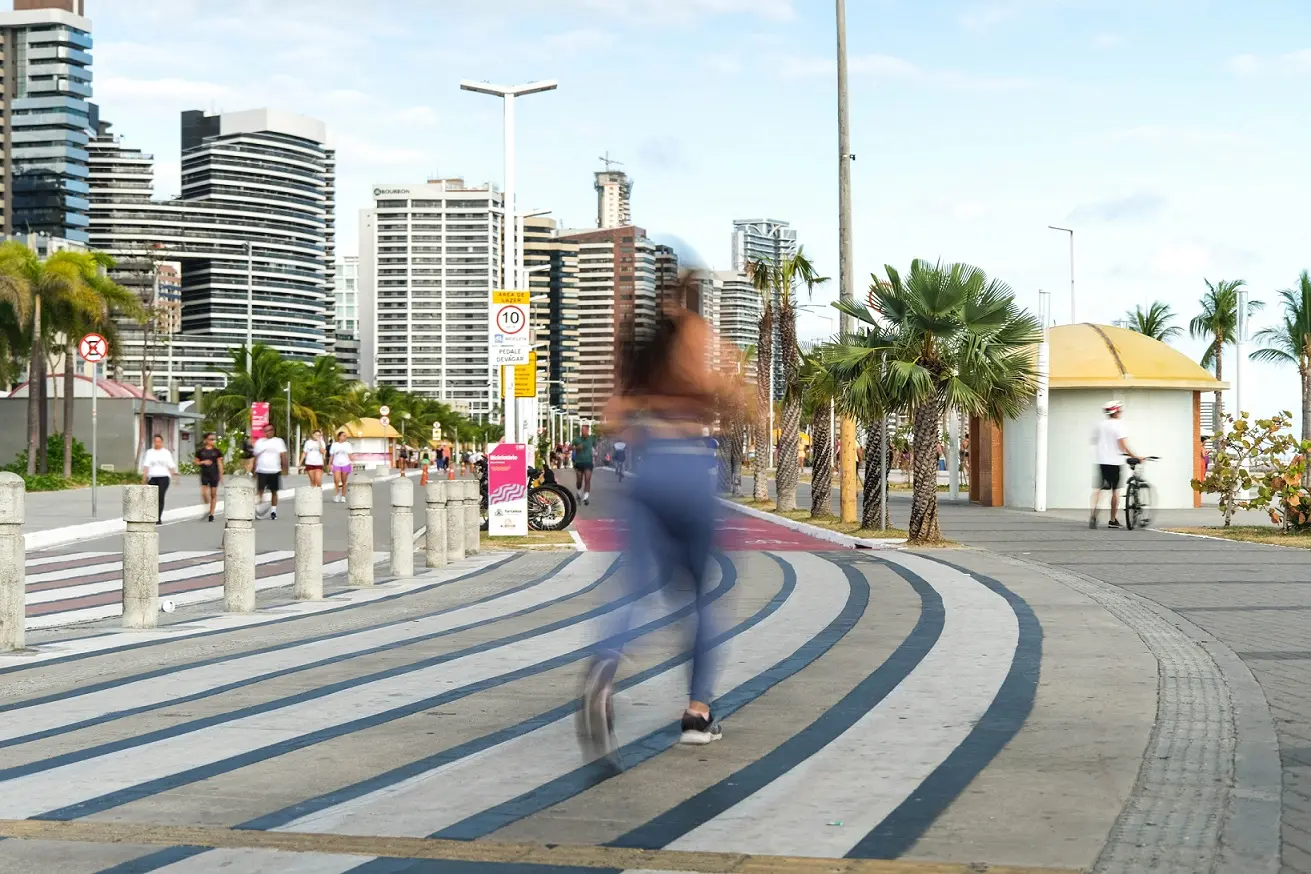 Foto mostra a orla urbana de Fortaleza, com prédios altos ao fundo e uma via exclusiva para pedestres e ciclistas. No centro, uma mulher corre pela calçada com desenho ondulado, enquanto outras pessoas caminham, andam de bicicleta e aproveitam o espaço público à beira-mar.