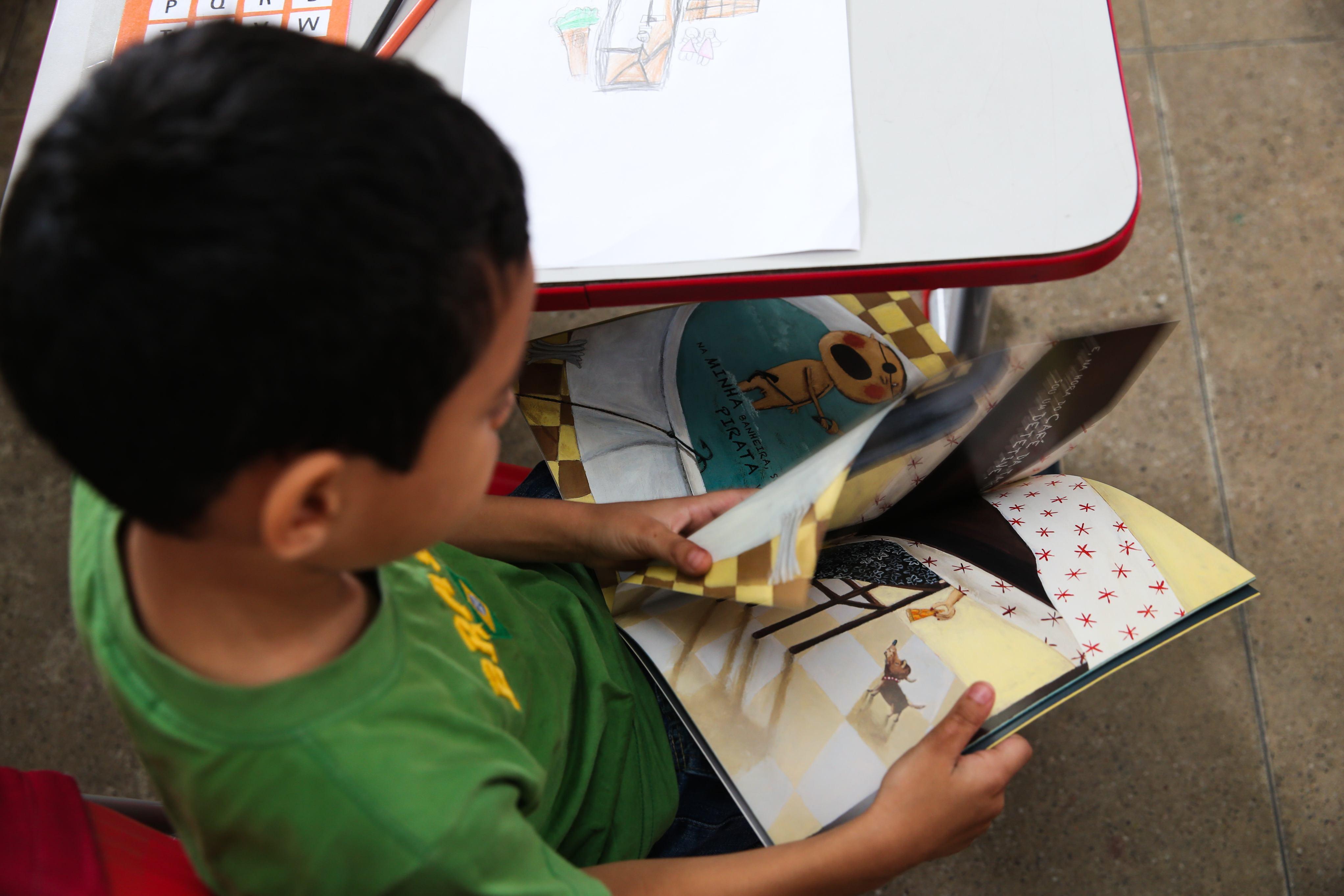 A foto mostra um menino com cabelo escuro e camisa verde está sentado a uma mesa, de costas para o observador. Ele está absorto na leitura de um livro ilustrado com páginas coloridas, que ele segura aberto à sua frente. As páginas do livro mostram desenhos e texto. Uma mesa escolar branca e vermelha está visível à esquerda, com papéis e alguns objetos de escrita em cima. O chão de azulejos cinzas é visível na parte inferior da imagem.