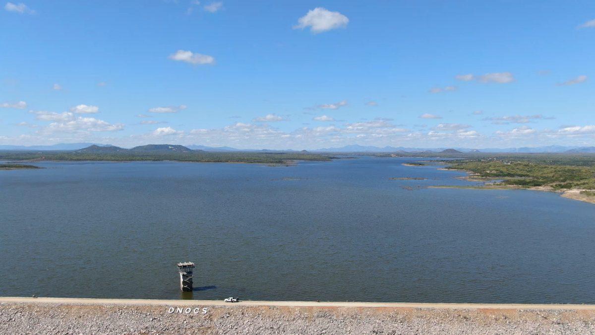 Foto do Açude Edson Queiroz, em Santa Quitéria, cheio de água, com parede de proteção com letreiro do DNOCS