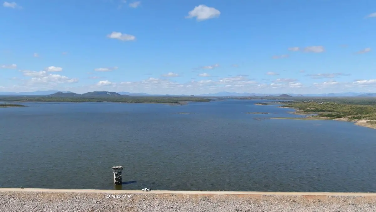 Foto do Açude Edson Queiroz, em Santa Quitéria, cheio de água, com parede de proteção com letreiro do DNOCS