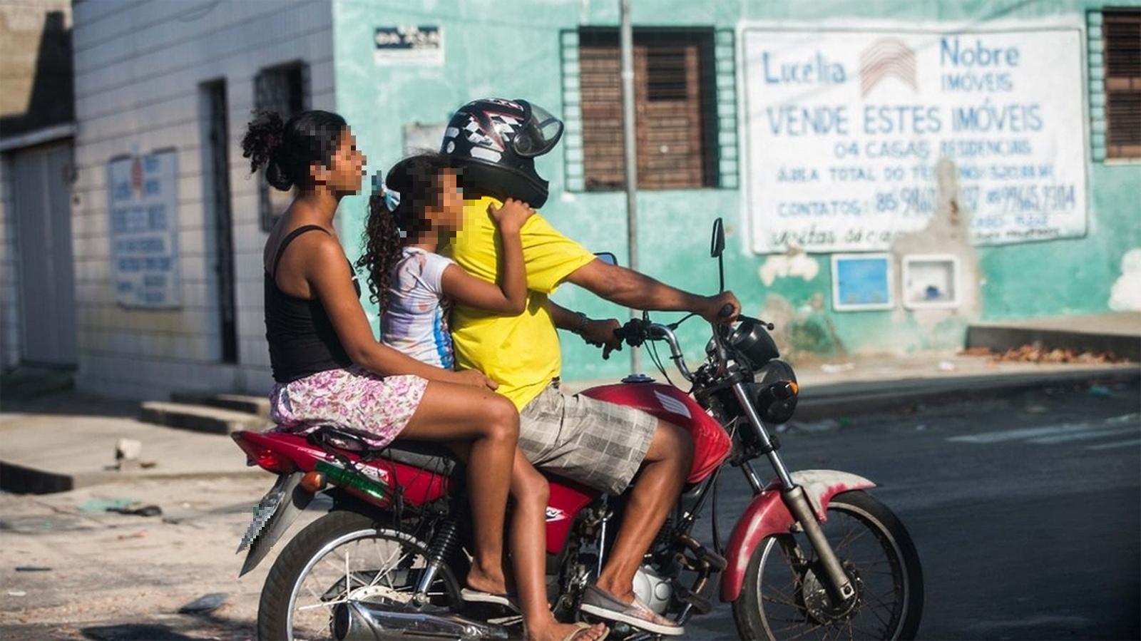 Imagem mostra três pessoas em cima de uma moto vermelha em rua do Ceará. O piloto é um homem de camisa amarela e bermuda xadrez cinza. No meio há uma criança, menina, de cabelo preso com um laço azul. Na garupa, uma mulher com camisa preta e short estampado.