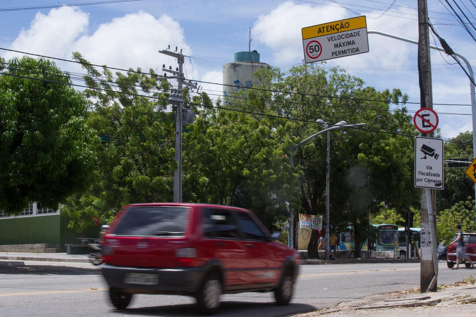 Imagem mostra carro Fiat Uno vermelho em avenida de Fortaleza. Ao lado, um poste com o aviso de que a via é fiscalizada por câmeras. Acima, uma placa indicando que a velocidade máxima permitida na via é de 50 km/h