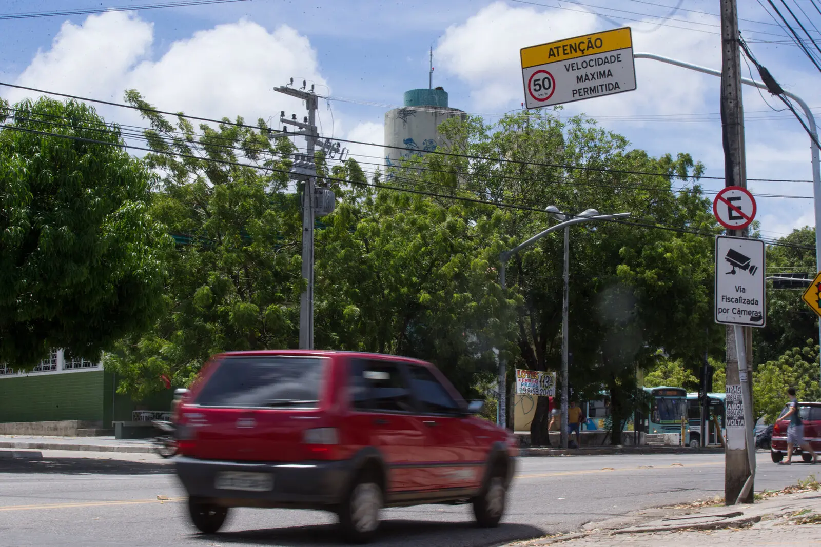 Imagem mostra carro Fiat Uno vermelho em avenida de Fortaleza. Ao lado, um poste com o aviso de que a via é fiscalizada por câmeras. Acima, uma placa indicando que a velocidade máxima permitida na via é de 50 km/h