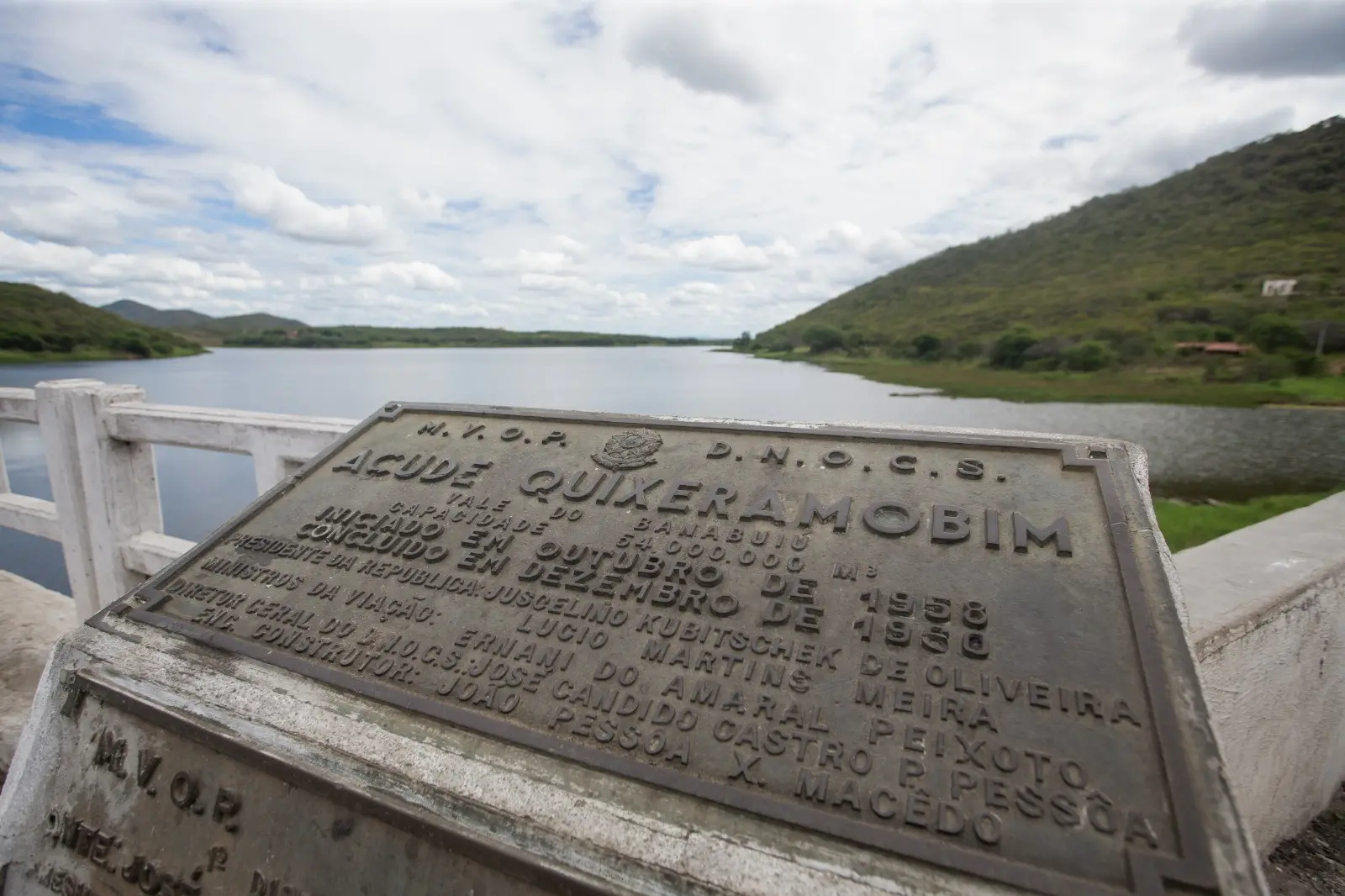 Foto que contém placa de inauguração do Açude Quixeramobim. Ao fundo, o açude do município