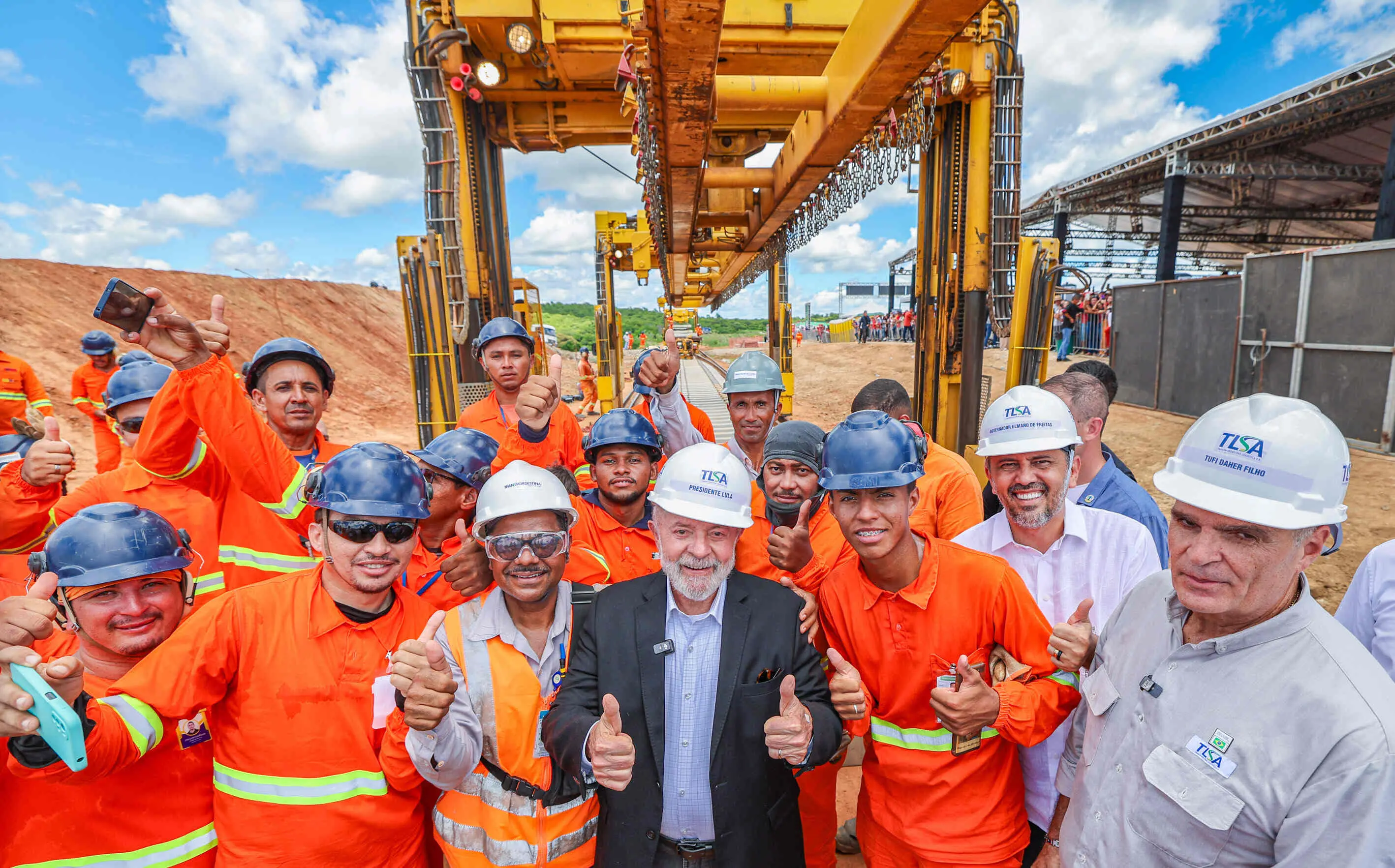Grupo de trabalhadores da construção civil posa para foto sorrindo e fazendo sinal de positivo com os polegares levantados. Todos vestem uniformes laranja com faixas refletivas e capacetes de segurança. Ao centro, Lula, de terno escuro e capacete branco com o nome “PRESIDENTE LULA”, também sorri e faz o gesto de positivo. Ao fundo, há uma grande estrutura metálica amarela utilizada em obras, trilhos, e um canteiro de obras com céu azul e nuvens. A cena transmite um clima de otimismo e celebração em visita institucional a uma obra de infraestrutura.