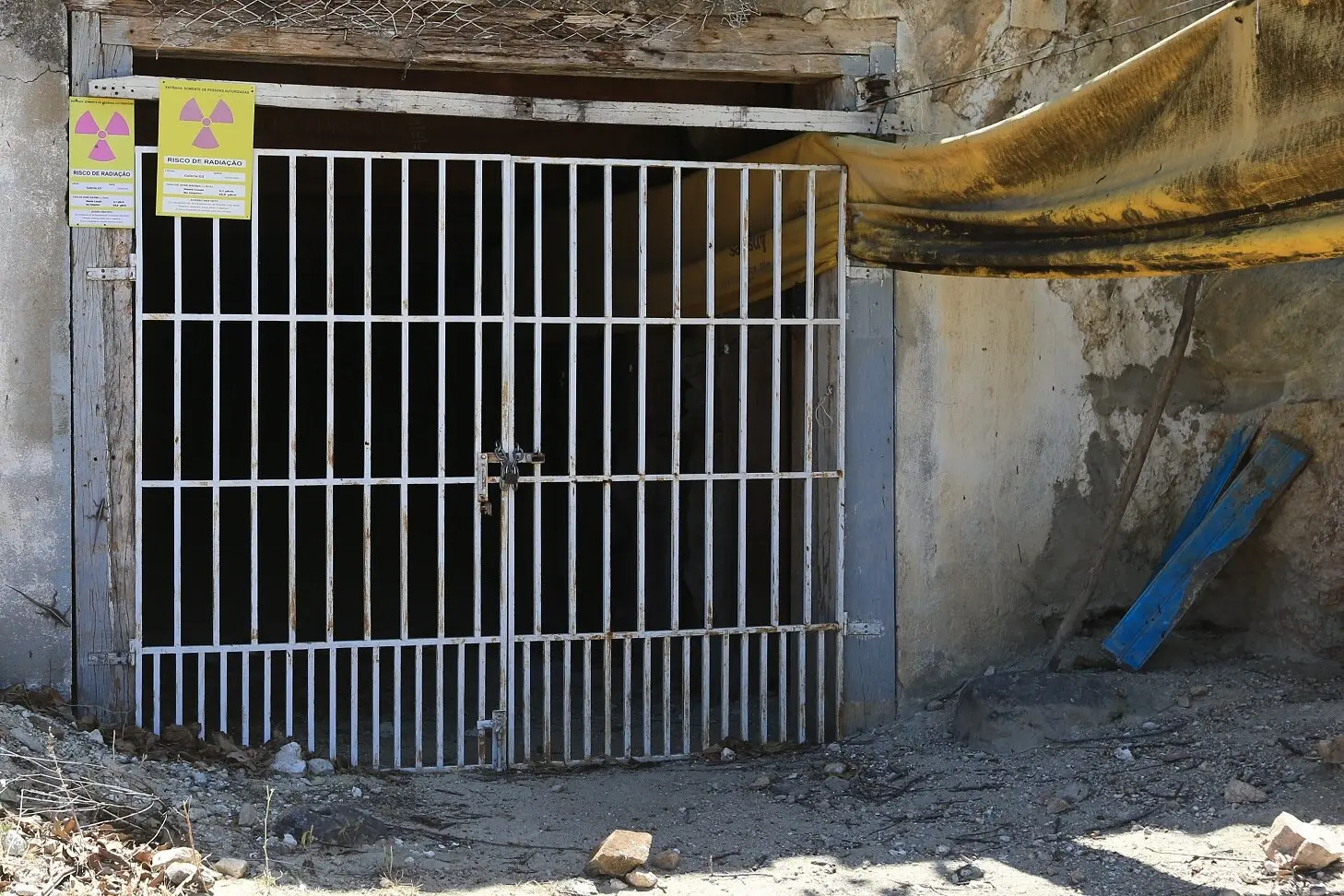 Foto de entrada de mina fechada com grades de ferro, contendo placas de alerta de radiação fixadas acima do portão.