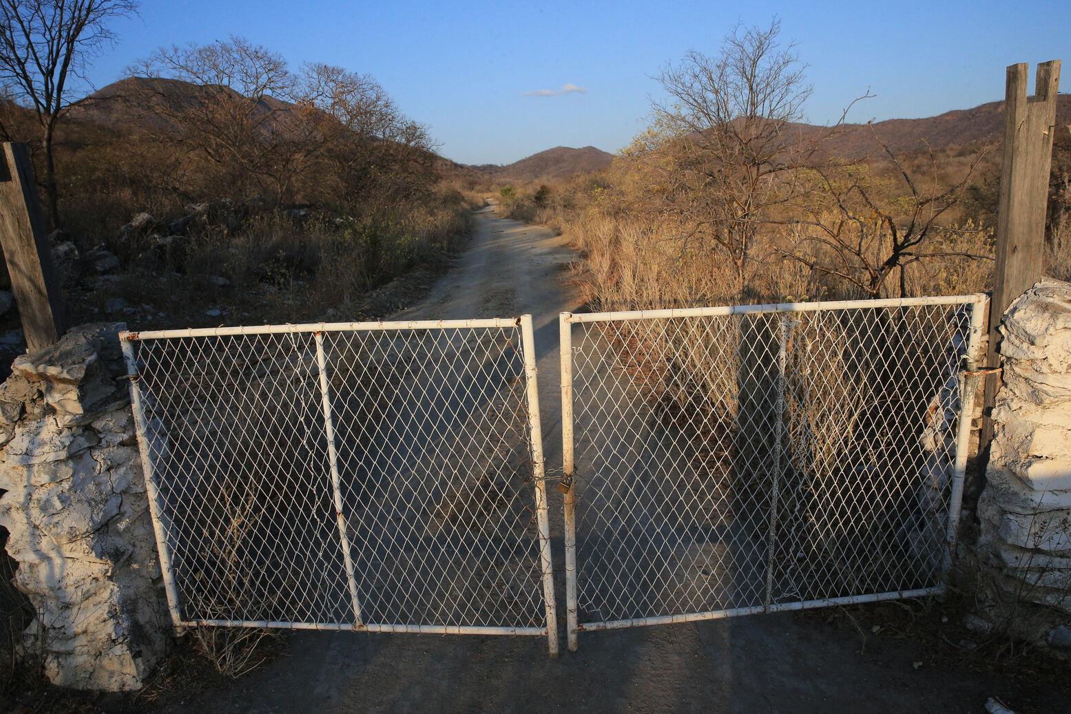 Foto de estrada de terra cercada por vegetação seca e portão metálico simples branco delimitando o acesso à área da mina.