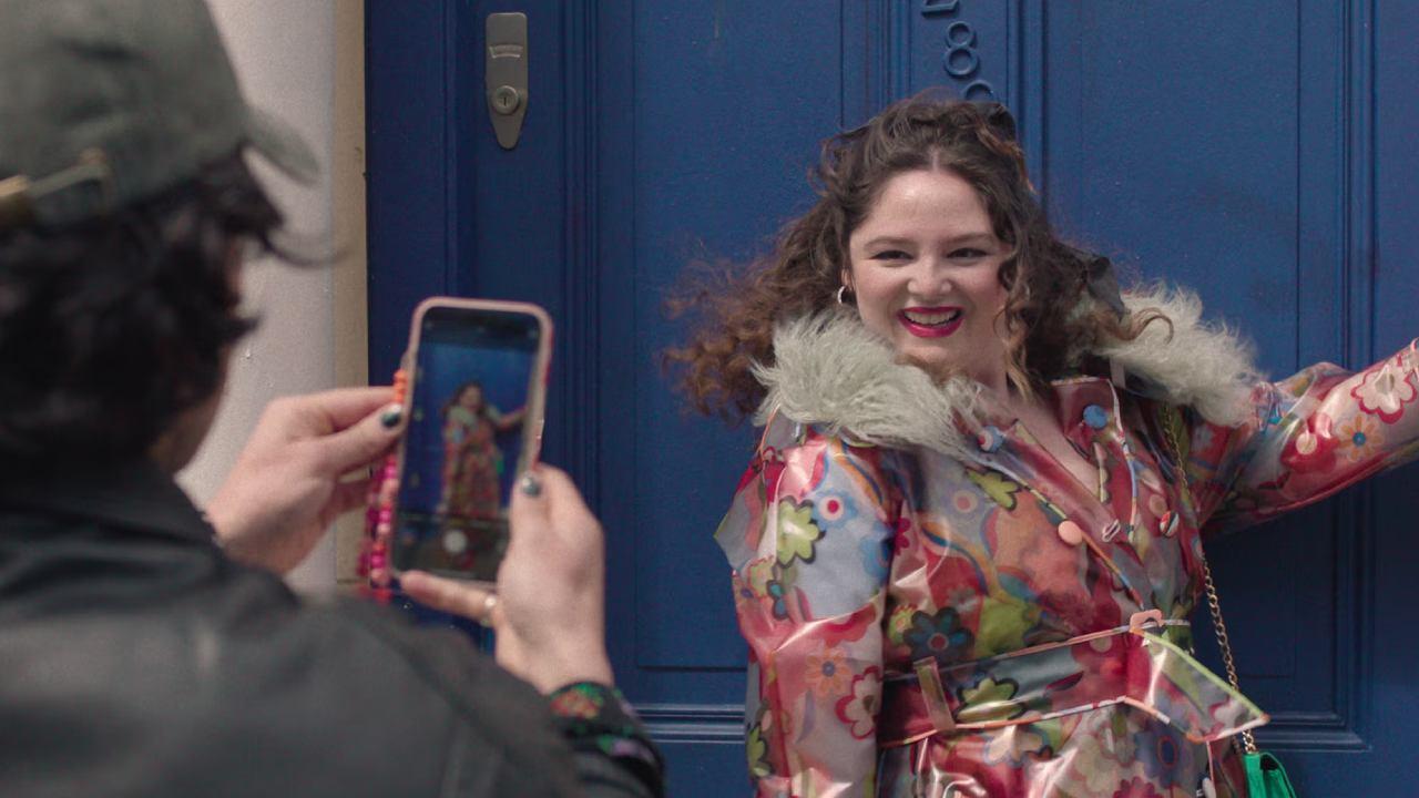 Atriz Megan Stalter caracterizada como Jessica posa para foto com casaco colorido em frete a uma porta azul