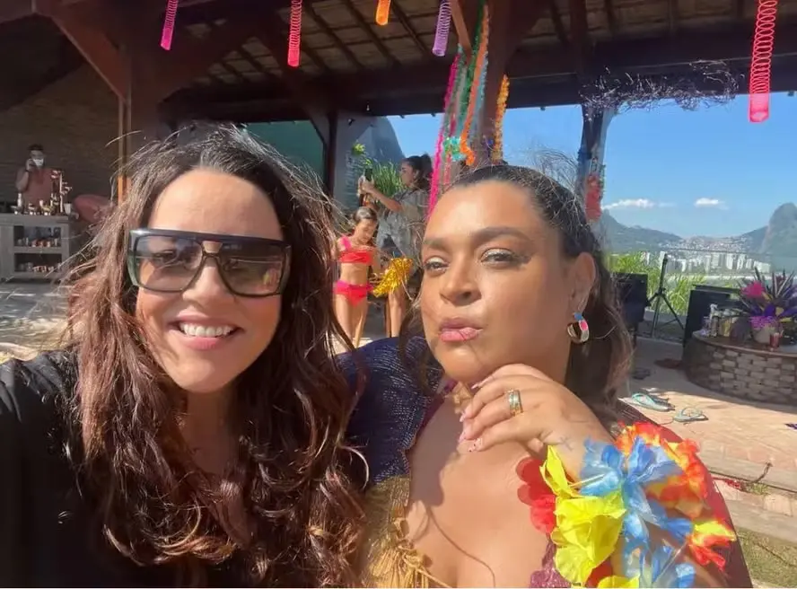 As cantoras Preta Gil e Ana Carolina sorrindo em uma festa ao ar livre com fundos de decoração colorida e pessoas ao fundo, celebrando um evento social.