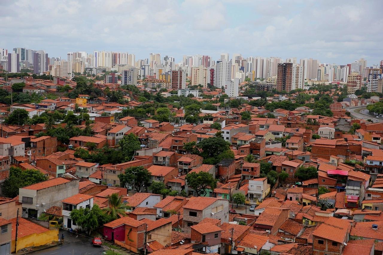 Foto com vista aérea de Fortaleza mostra forte contraste urbano, com área de casas populares e telhados vermelhos em primeiro plano e prédios altos ao fundo.