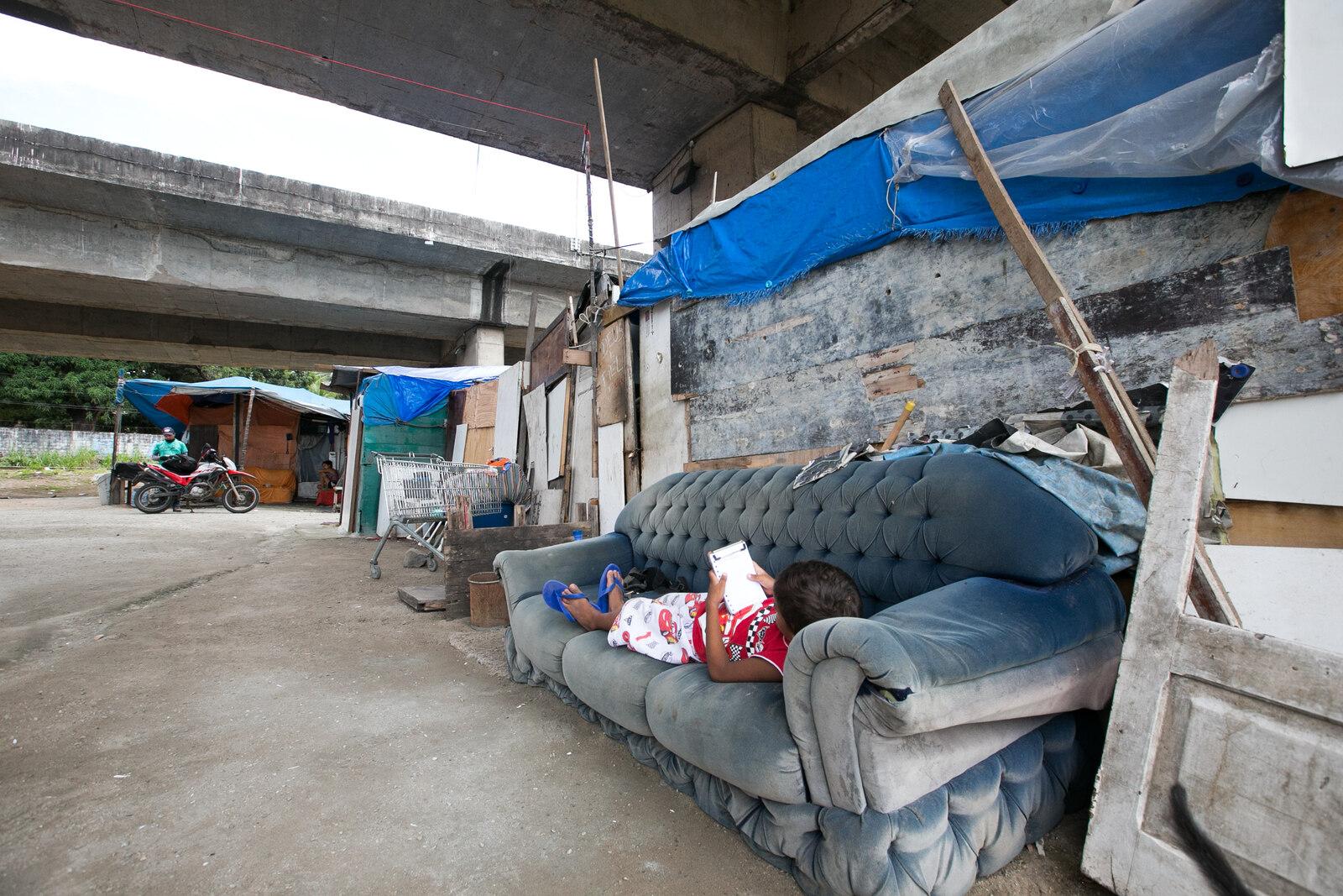 Criança mexe em celular deitada em sofá debaixo de viaduto