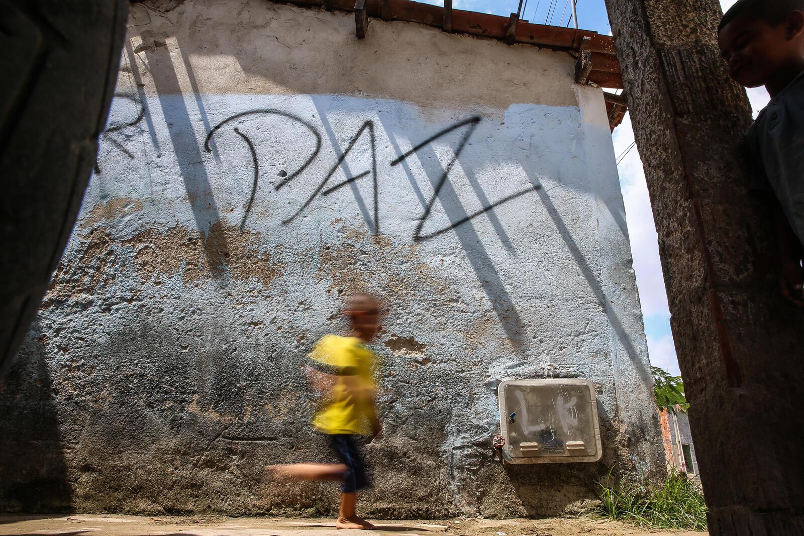 Menino com blusa amarela corre em frente a parede com o escrito Paz
