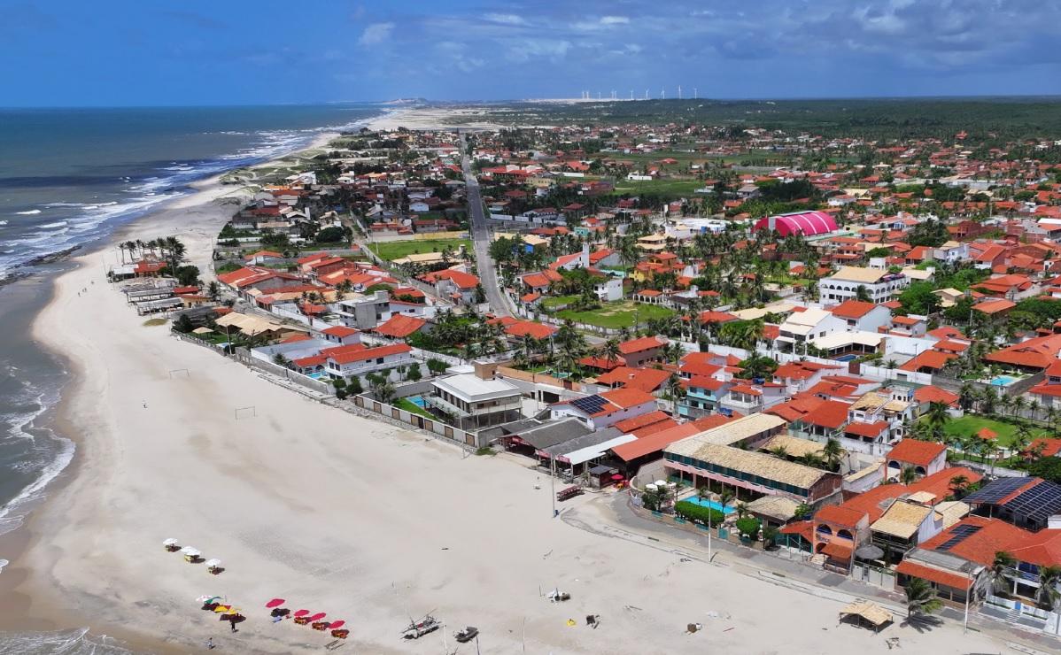 Vista aérea da Praia da Caponga, no litoral cearense, mostra uma faixa de areia clara, mar calmo e casas com telhados vermelhos, evidenciando ocupação voltada ao turismo e veraneio. Ao fundo, aparecem áreas verdes e aerogeradores.
